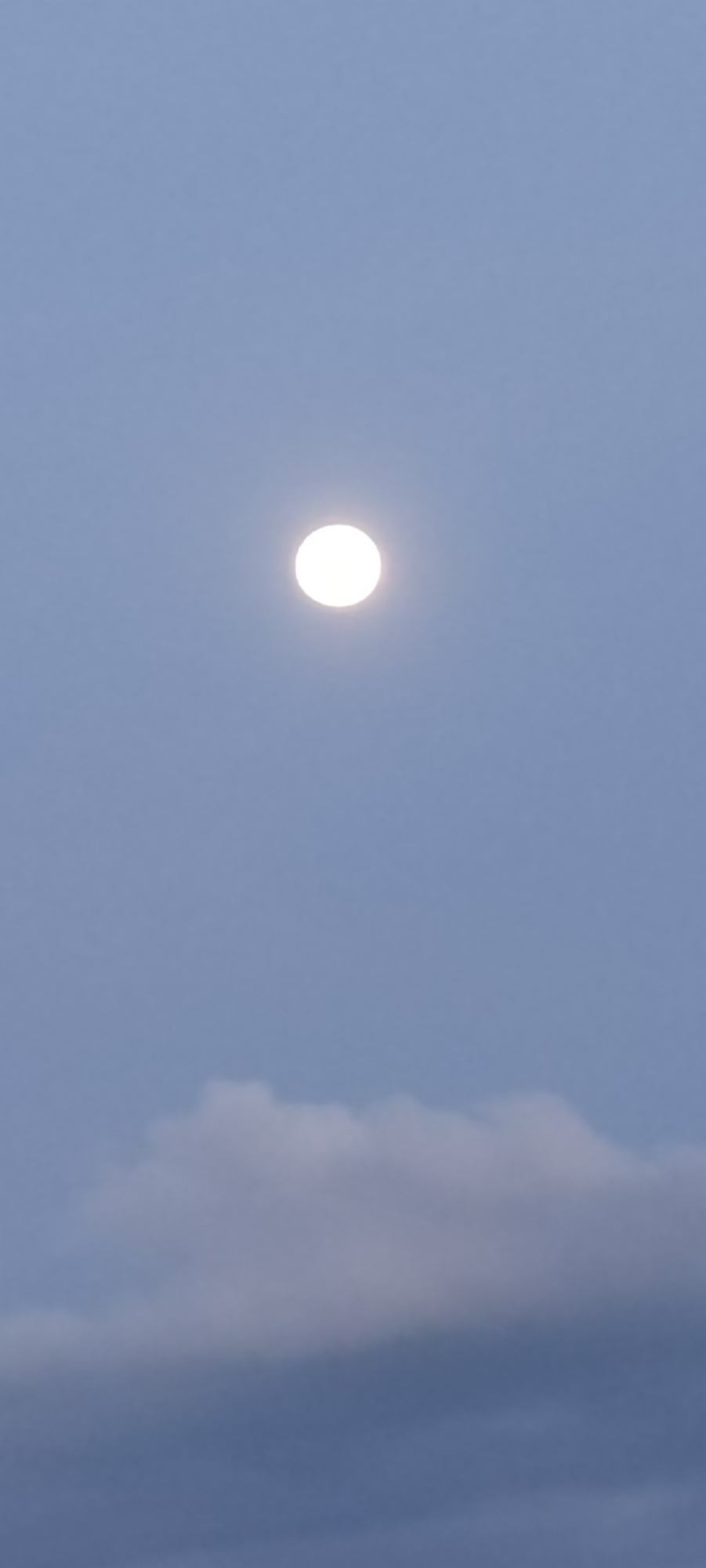 A bright, circular moon hangs in the blue sky. The moon is almost directly overhead, centrally located in the image. Soft, diffuse clouds are visible near the bottom of the frame, suggesting a slightly overcast condition. There is no indication of people, buildings, or any other features that would identify the location as Cotgapu, Indonesia, or any other specific city. The focus is on the sky and the moon.