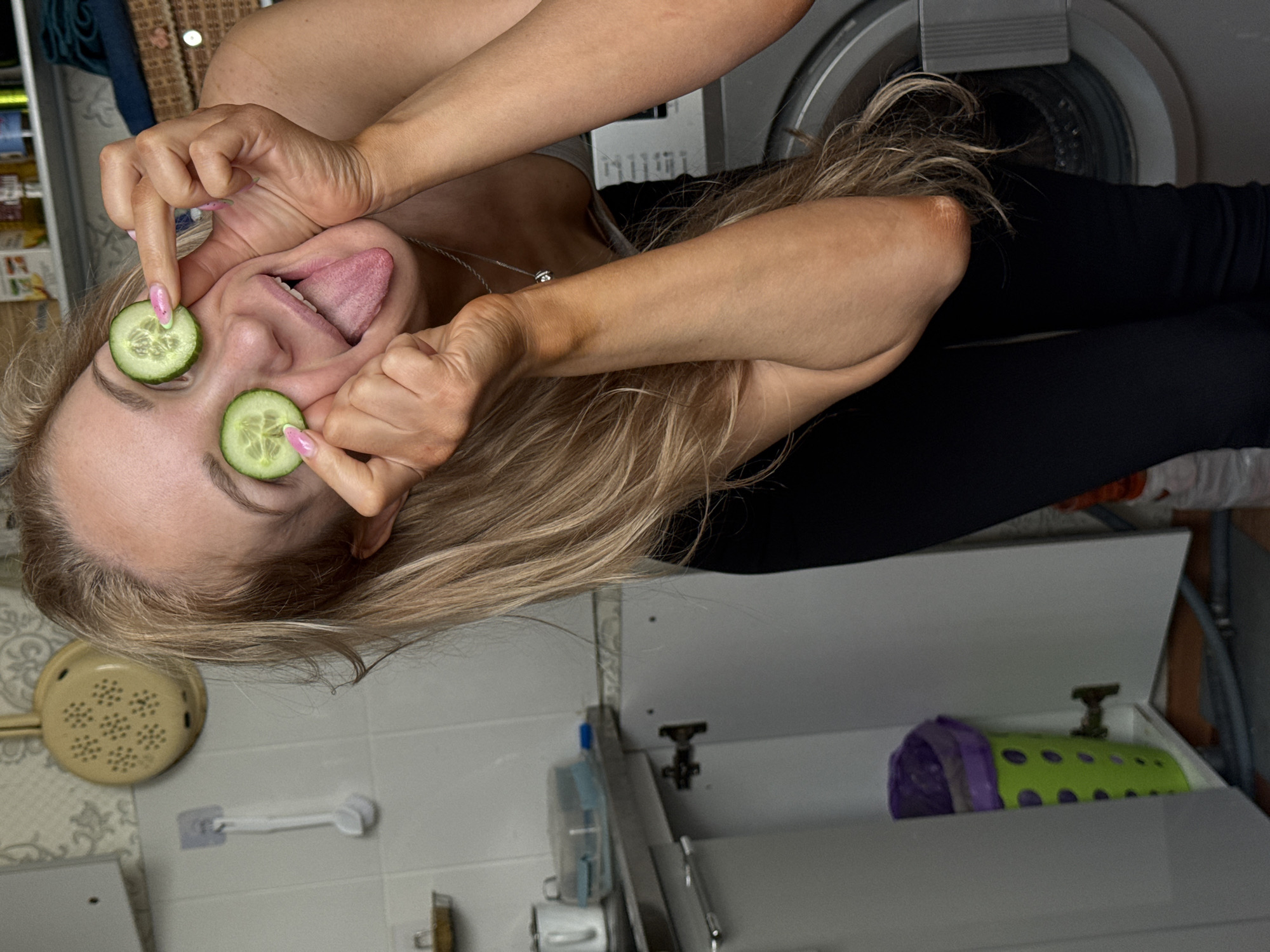 A woman with long blonde hair is in a bathroom with white tile and white cabinets. She is holding cucumber slices over her eyes while sticking her tongue out. Behind her is a washing machine. A colander is on the wall to the left. Inside one of the cabinets is a purple and green object. The image provides no information to indicate the location is Chaikovsky, Russia.
