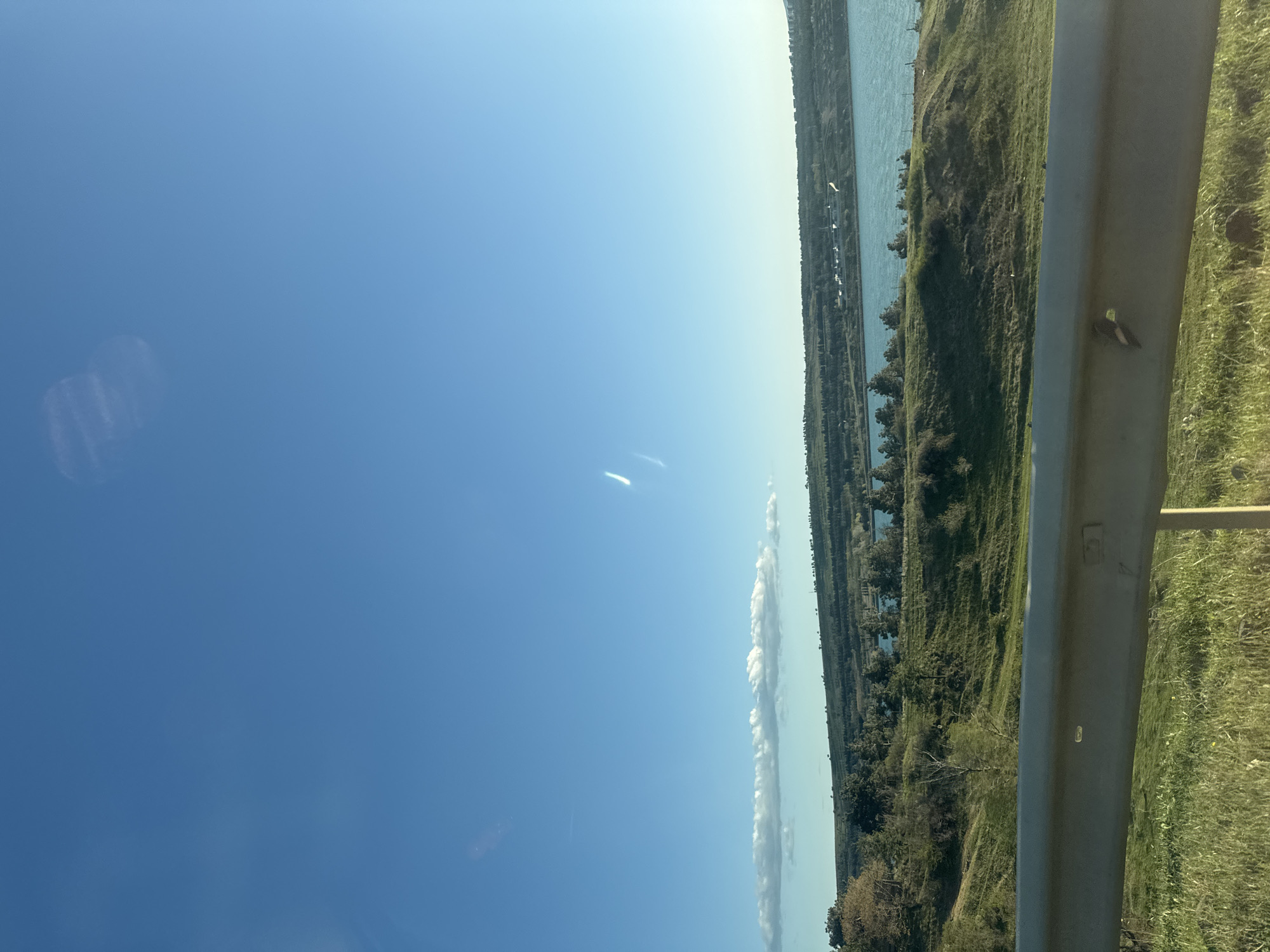 The image shows a view from a roadside in Tbilisi, Georgia. A blue sky fills the left side, with a narrow, vertical line of white clouds extending from the center-right upwards. To the right of the clouds is a waterway with tree-lined banks. A metal guardrail runs vertically along the right side of the image, with grassy vegetation behind it. Buildings, trees, and possibly other vegetation are visible in the distance, alongside the waterway.