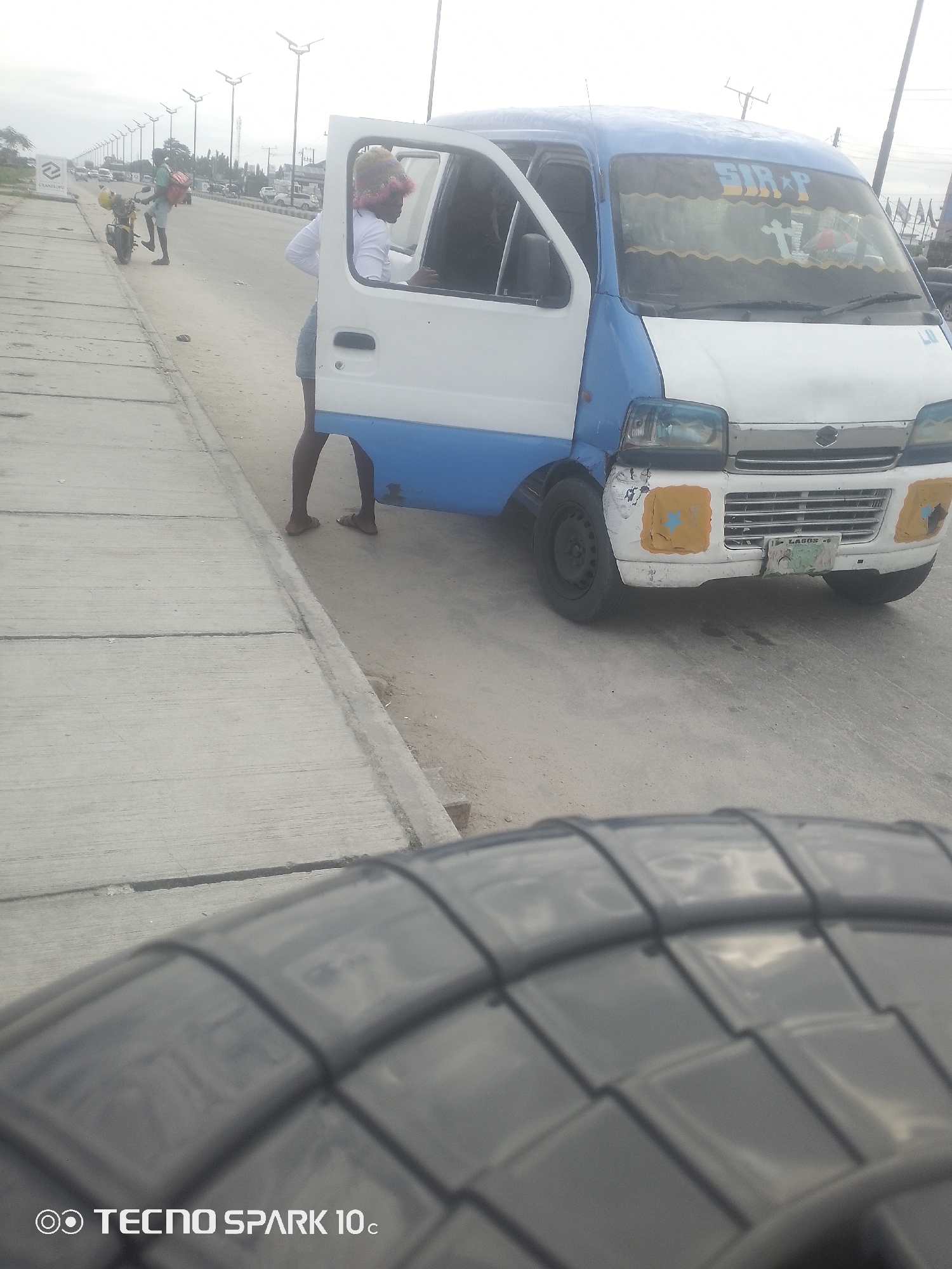 A white and blue minibus is parked on the side of a road in Lekki, Nigeria. The driver's side door of the minibus is open, and a person wearing a white long-sleeved shirt and denim shorts stands with their back to the camera, appearing to be interacting with someone inside the vehicle. A person on a motorcycle with a red bag on the back is also visible on the left side of the road, further down.
The scene appears to be set during the day, under an overcast sky, suggesting it might be a cloudy day. The road is paved with concrete on the left, and there's a sandy shoulder. Streetlights are visible in the background, lining the road. Other vehicles can be seen in the distance. The minibus itself shows signs of wear and tear, with some dents and patches on the front bumper.
Visible text on the minibus includes "SIR P" at the top of the windshield, and what appears to be a license plate that reads "LAGOS." There is also text at the bottom left of the image that says "O TECNO SPARK 10c." The overall impression is of a typical street scene in a bustling Nigerian city.
