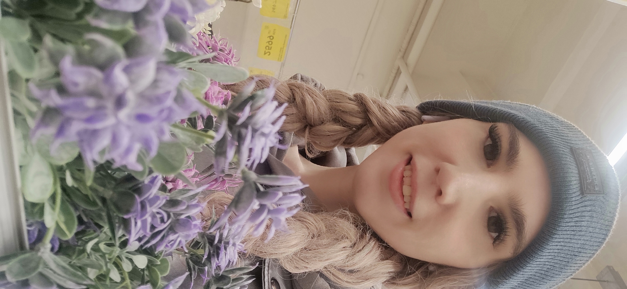 A woman with braided blonde hair and a blue beanie is positioned on the right side of the frame. Her smile is visible. To the left of the woman are artificial purple flowers, some of which are out of focus. In the background, partially visible, are some yellow stickers. The location is the city of Chaikovsky, Russia.