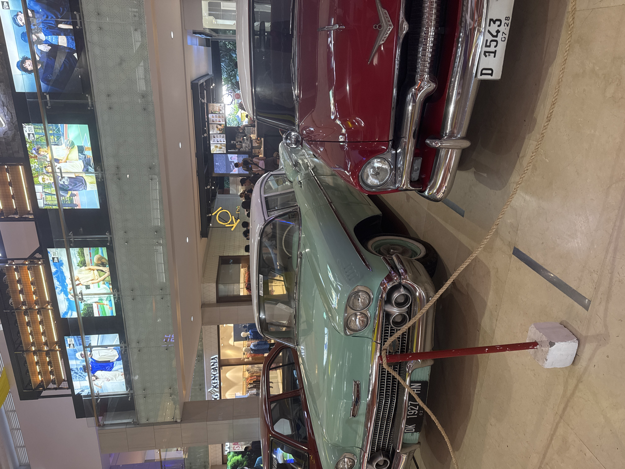 The scene features two vintage cars, one red and one teal, displayed within a building, likely a shopping mall in an unknown city, unknown. The cars are positioned next to each other, partially overlapping in the frame. The red car has a license plate that reads "D 1543", with "G7.28" below. The teal car's license plate reads "DK 1927 HN", with "02" below. A rope barrier, supported by a red pole and white concrete block, surrounds the teal car.
In the background, a multistory atrium can be observed. Digital screens showing various advertisements are attached to the walls. There are shops with glass fronts and mannequins. People are visible in the background. The floor is a light beige color.
