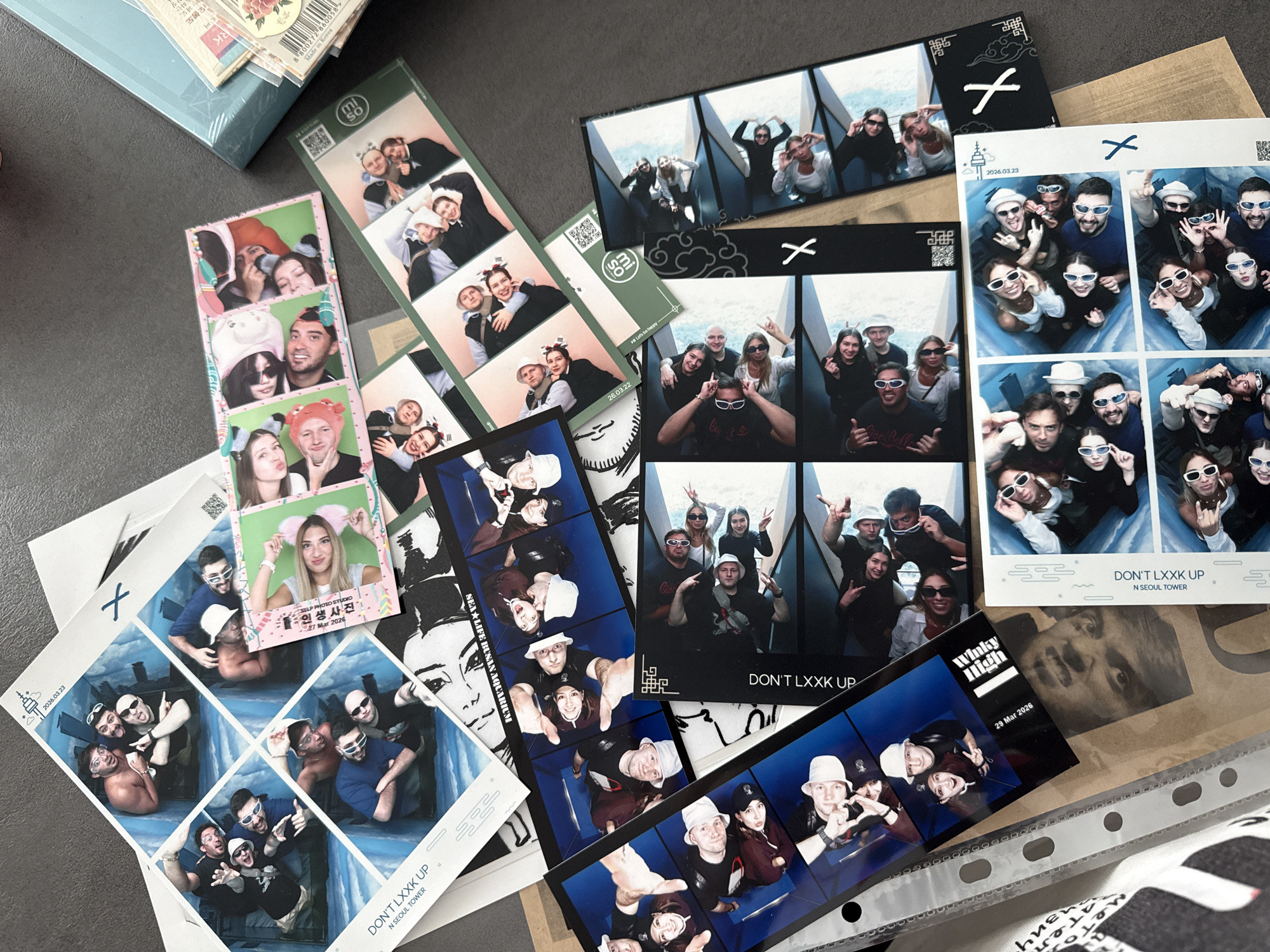 The image shows a collection of photo booth strips scattered on a gray surface, seemingly a tabletop. The strips vary in color, from black and white to full-color, and depict groups of people in playful poses. Some strips include dates, such as "2026.03.23," "27 Mar 2026," "26.03.22," and "29 Mar 2026." The text "DON'T LXXK UP" is printed on multiple strips, along with "N SEOUL TOWER," indicating the location of the photo booth within the Seoul Tower. Other text includes "SELP PHOTO STUDIO 인생사진," "SEA LIFE BUSAN AQUARIUM," and "Winky High." One strip has Korean text and the phrase "Let's be happy." A box with packaging labels is visible in the top left corner. A clear plastic sheet with metal binding can be seen in the bottom right, along with what appears to be a fabric with text.