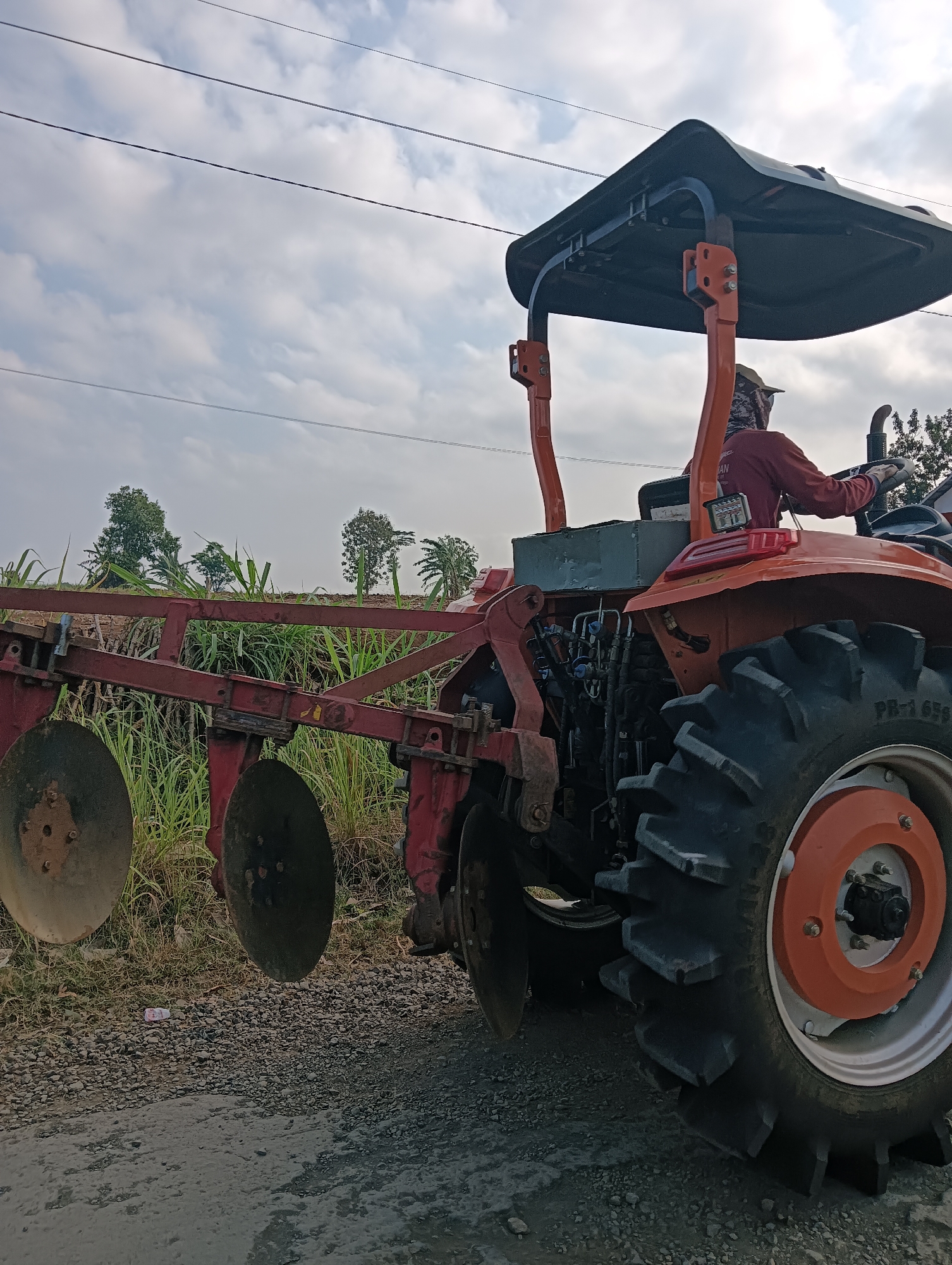 An orange tractor with a disc plow attachment is parked on a dirt road. A person wearing a red long-sleeved shirt, a hat, and a patterned mask is sitting in the driver's seat. The tractor is facing away from the viewer and slightly to the right. In the background, there are green fields with trees and a cloudy sky. The scene appears to be in Cluwak, Indonesia.
