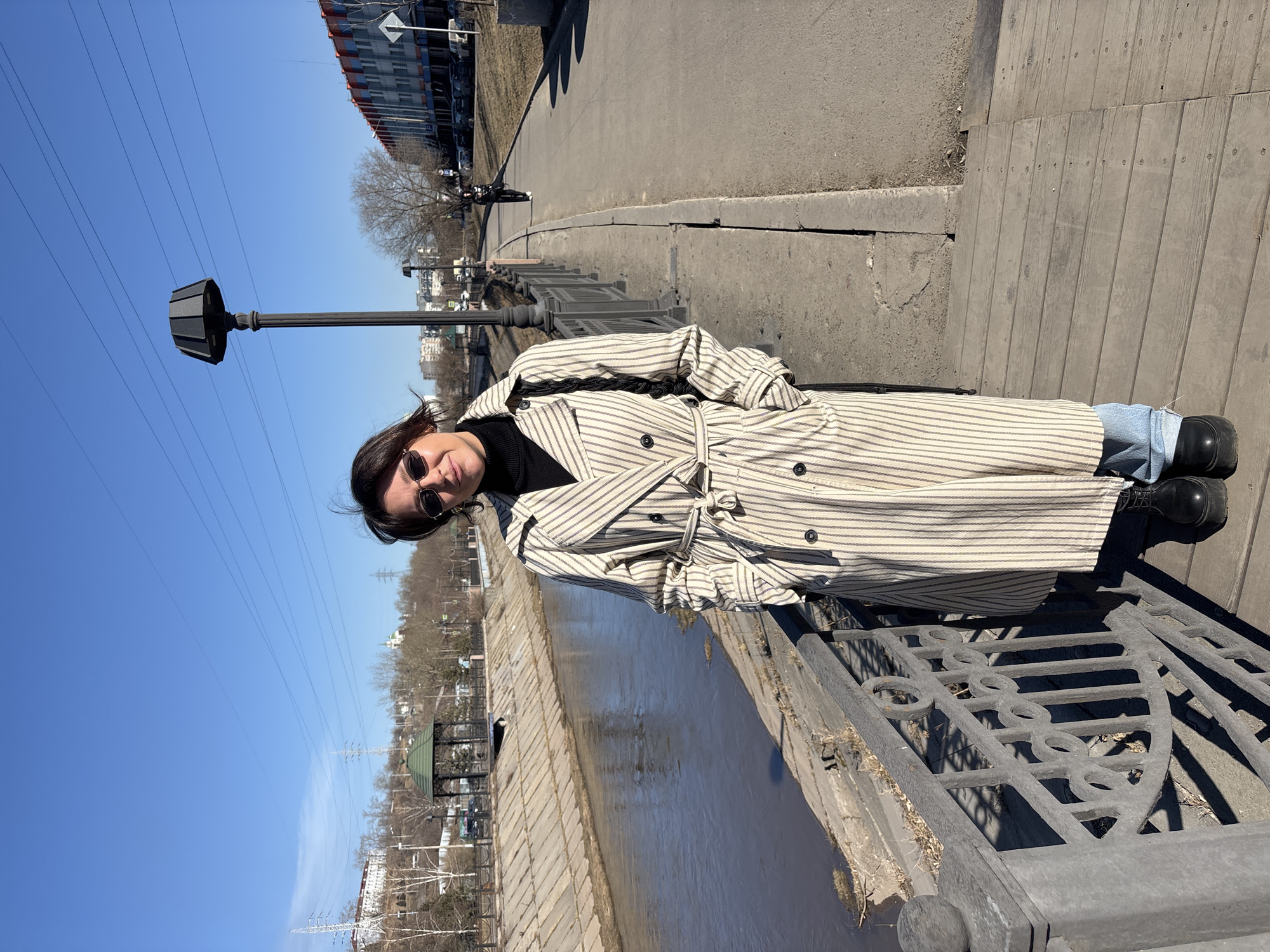 A woman wearing a striped trench coat, black turtleneck sweater, blue jeans, and sunglasses stands on a bridge. She leans against the railing on the right side of the image. Behind her, the water reflects the sky and nearby foliage. The bridge has a metal railing with decorative circles. In the background, there is a park with trees, a gazebo-like structure, and buildings. A lamppost stands to the left of the woman. Power lines are visible in the sky. The photograph appears to be taken on a sunny day. The location is likely in the city of Krasnoyarsk, Russia.