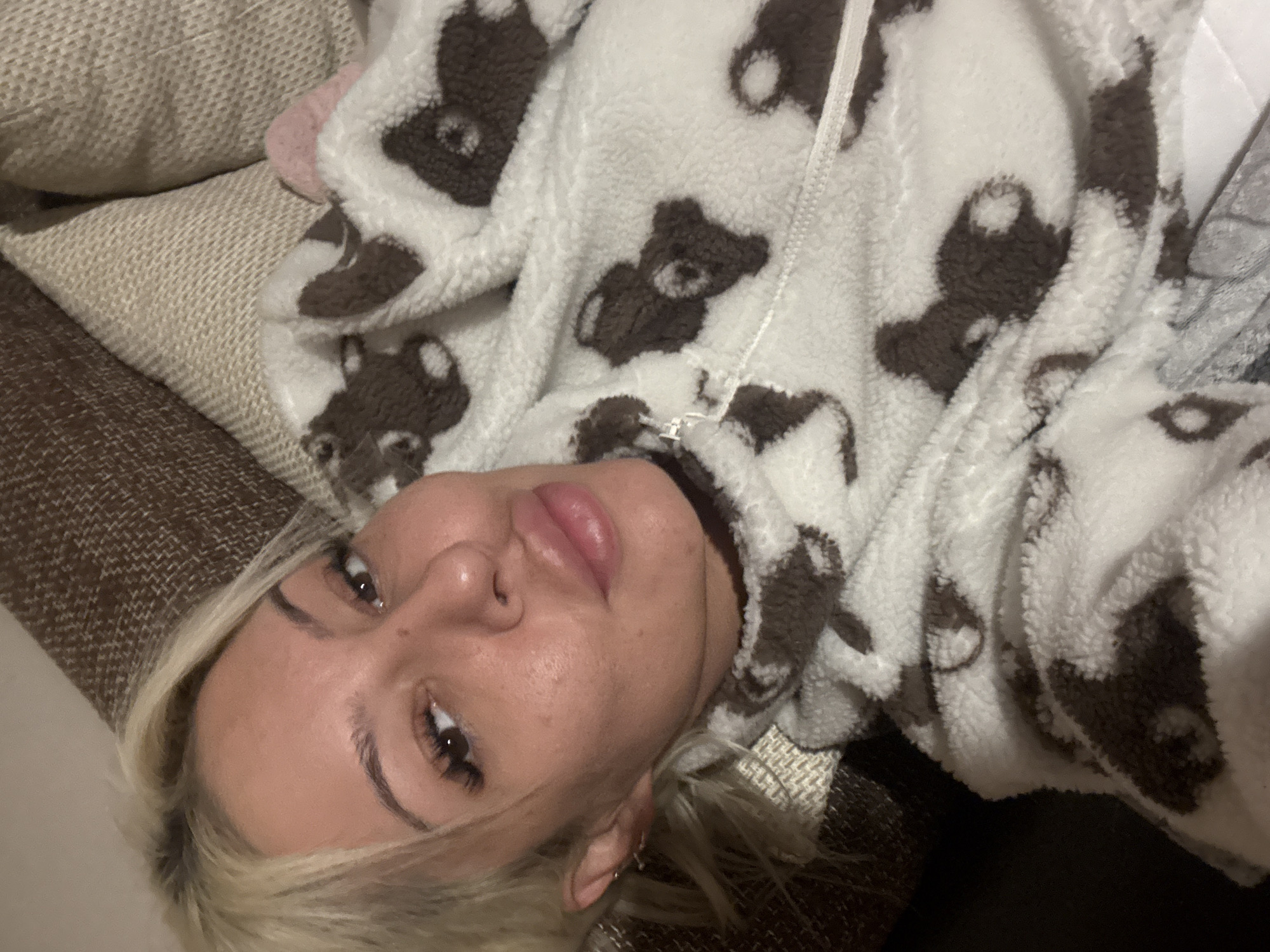 A person is depicted in a close-up, slightly overhead shot, lying down indoors. The subject is a young adult with blonde hair, dark eyes, and a neutral to slightly pouting expression on their face. They appear to be wearing light makeup, including defined eyebrows and long eyelashes, and possibly lip gloss.
The person is dressed in a cozy, plush white garment, likely a onesie or robe, covered in a repeating pattern of brown teddy bears. A zipper is visible down the front of the garment. They are positioned on what appears to be a textured brown and beige surface, possibly a sofa or bed, with soft furnishings or blankets around them. The composition suggests a casual, relaxed indoor setting, potentially a home in Plovdiv, Bulgaria. The lighting is soft and even, consistent with an indoor environment. No text is visible in the image.