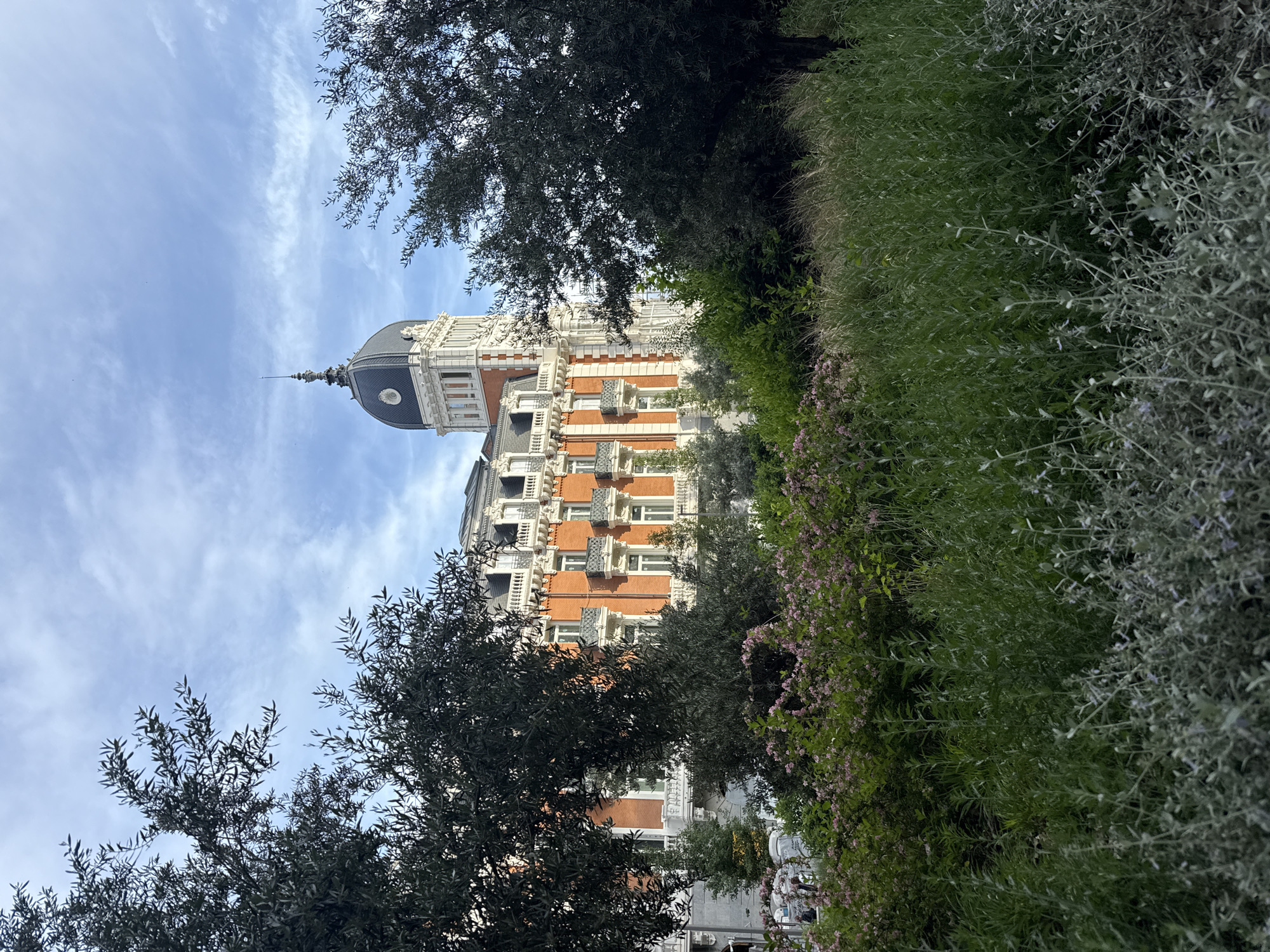 The image shows a section of the exterior of the Palacio de Cibeles in Madrid, Spain, viewed from a slightly low angle. The building's facade is characterized by red brick and white decorative elements. Its dome rises into a blue sky with scattered clouds. Trees and greenery partially obscure the base of the building.