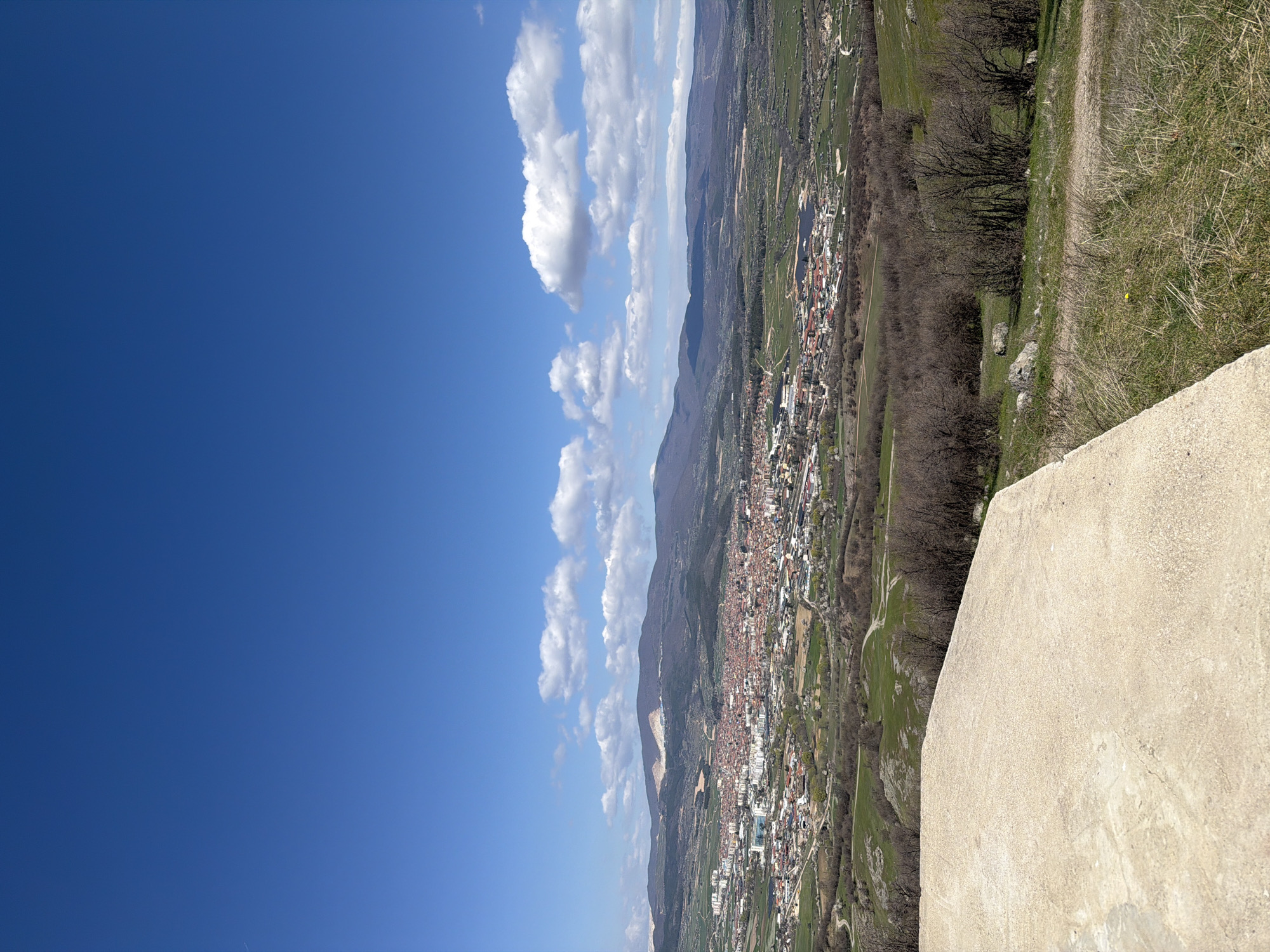 An elevated, oblique perspective of a city, presumably Bata, Bulgaria, is presented under a clear, blue sky interspersed with cumulus clouds. The city itself is densely populated with buildings and appears to be nestled in a valley, surrounded by hills and sparsely vegetated areas. A concrete wall occupies the foreground, adding to the sense of height. Bare trees line the landscape, suggesting a season with less foliage, possibly early spring or late autumn. A dirt path can be seen near the right edge, adjacent to grassy terrain.