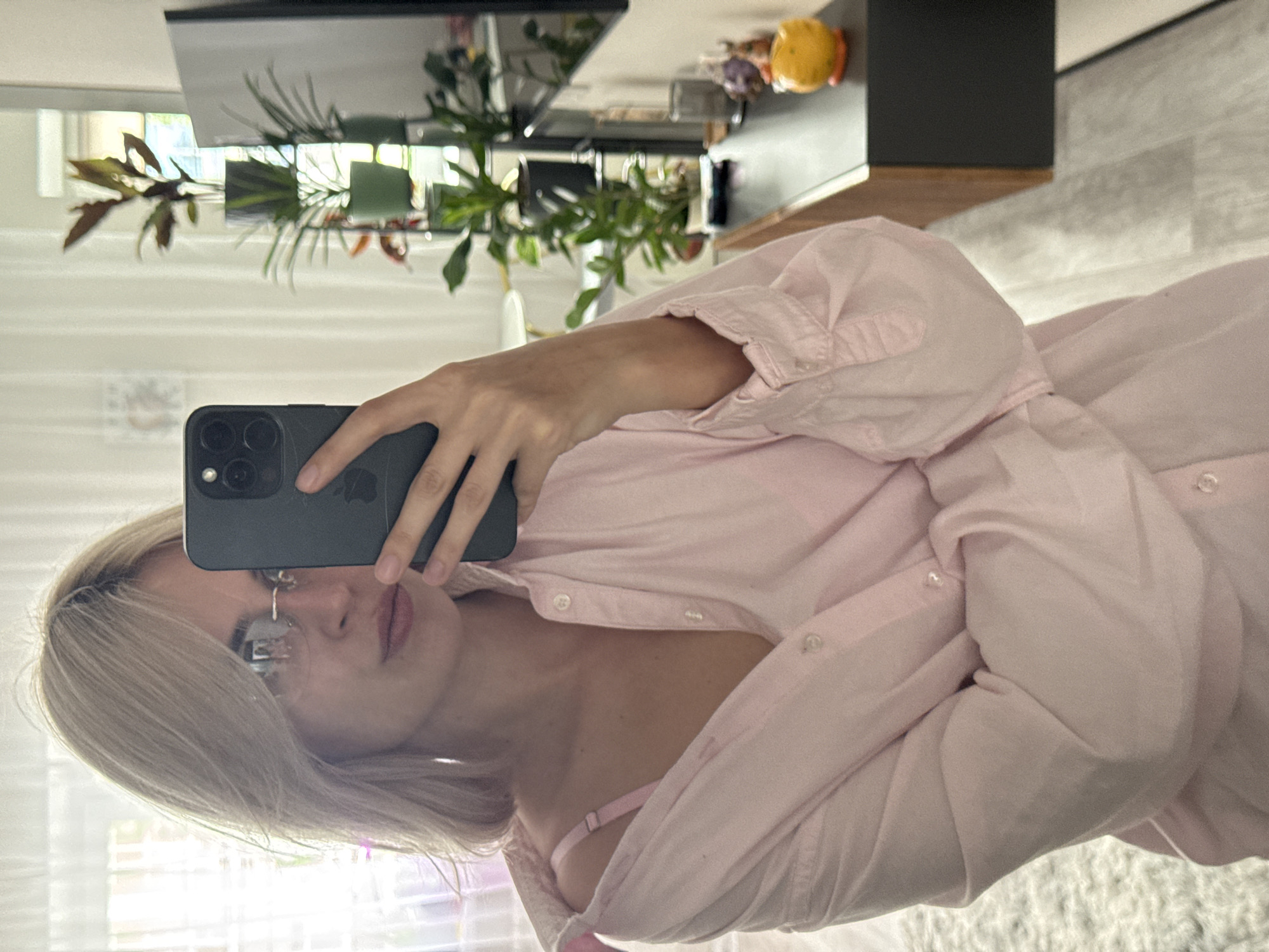 A person is taking a selfie in a mirrored reflection. The person has blonde hair, wears glasses and a light pink button-down shirt that is slightly unbuttoned revealing a pink bra strap. They are holding a dark-colored smartphone to take the picture, obscuring their face. The background visible through the mirror reflects a living room-like space with a television, a plant, and a yellow spherical object on a table. The location appears to be an indoor space in Saratov, Russia based on geographical analysis and cross-referencing of architectural features.