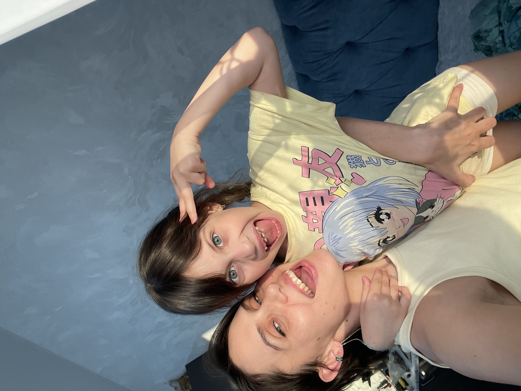 This media file presents a joyful indoor scene featuring two females, likely an adult and a child, positioned side-by-side and looking directly up at the camera. The image is taken from an overhead perspective, with their heads towards the bottom right and bodies extending towards the top left.
Both individuals have dark hair and light-colored eyes (appearing blue or green) and are smiling broadly with their mouths open, conveying happiness and playfulness. The adult, positioned on the right, wears a light-colored (possibly cream or off-white) tank top and a thin silver necklace. A distinctive green-banded earring is visible on her left ear. The child, on the left, wears a bright yellow t-shirt adorned with anime-style graphics featuring a blue-haired character and a cat, along with some visible Japanese text. She is making a "rock on" or similar playful hand gesture with her left hand, with her index and pinky fingers extended upwards. A dental appliance is visible in her open mouth.
The setting appears to be a domestic interior. A textured, greyish-blue wall forms the background on the left. On the right and top, a dark blue, possibly upholstered piece of furniture or bedding is visible. Bright natural light streams in from an unseen window, casting strong, defined shadows on their arms and clothing, indicating a sunny day, likely morning or afternoon. The overall mood is one of warmth and candid amusement. The provided context indicates the location is Izhevsk, Russia.