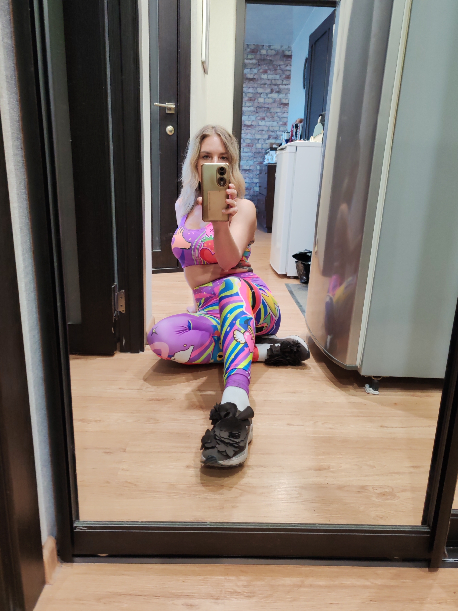 A fair-skinned woman with blonde hair is taking a selfie in what appears to be an apartment or house. She is kneeling on a light-colored wooden floor, wearing a vibrant purple, pink, and blue athletic outfit consisting of a sports bra and leggings. Her outfit features cartoon-like designs. She is holding a smartphone in her hand, presumably to take the picture, and her face is partially obscured by the phone.
She is wearing dark-colored shoes with white socks. The setting is indoors, and the background shows a door on the left, another door leading to what seems to be a kitchen or dining area with a refrigerator visible on the right. There are elements suggesting a residential interior, like the fridge and the style of the flooring. The location in the city of Petropavlovsk-Kamchatsky, Russia cannot be determined.