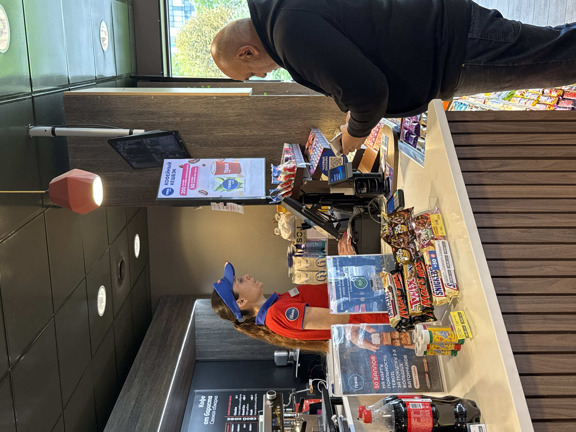 This image depicts the interior of a TEBOIL service station convenience store, likely located in Saint Petersburg, Russia, judging by the Russian language signage. A female cashier, wearing a red uniform shirt and a blue visor, stands behind a counter. She has dark hair pulled back in a ponytail and is looking towards a male customer. The male customer, with a bald head and beard, is facing away from the camera and is interacting with a handheld scanner at the counter.
On the counter, various items are displayed, including bottles of dark soda, candy bars such as Snickers, Mars, and Twix, and small packaged goods. Behind the cashier, shelves are visible with more packaged items, including what appear to be coffee bags. Above the counter, a digital screen displays promotional information, and a red geometric light fixture hangs from the ceiling. The environment is well-lit, with a combination of overhead lights and lighting from display shelves. The counter itself is white with a wooden paneled base. In the background, a window shows greenery, suggesting an outdoor area. The overall scene is typical of a modern convenience store at a gas station.