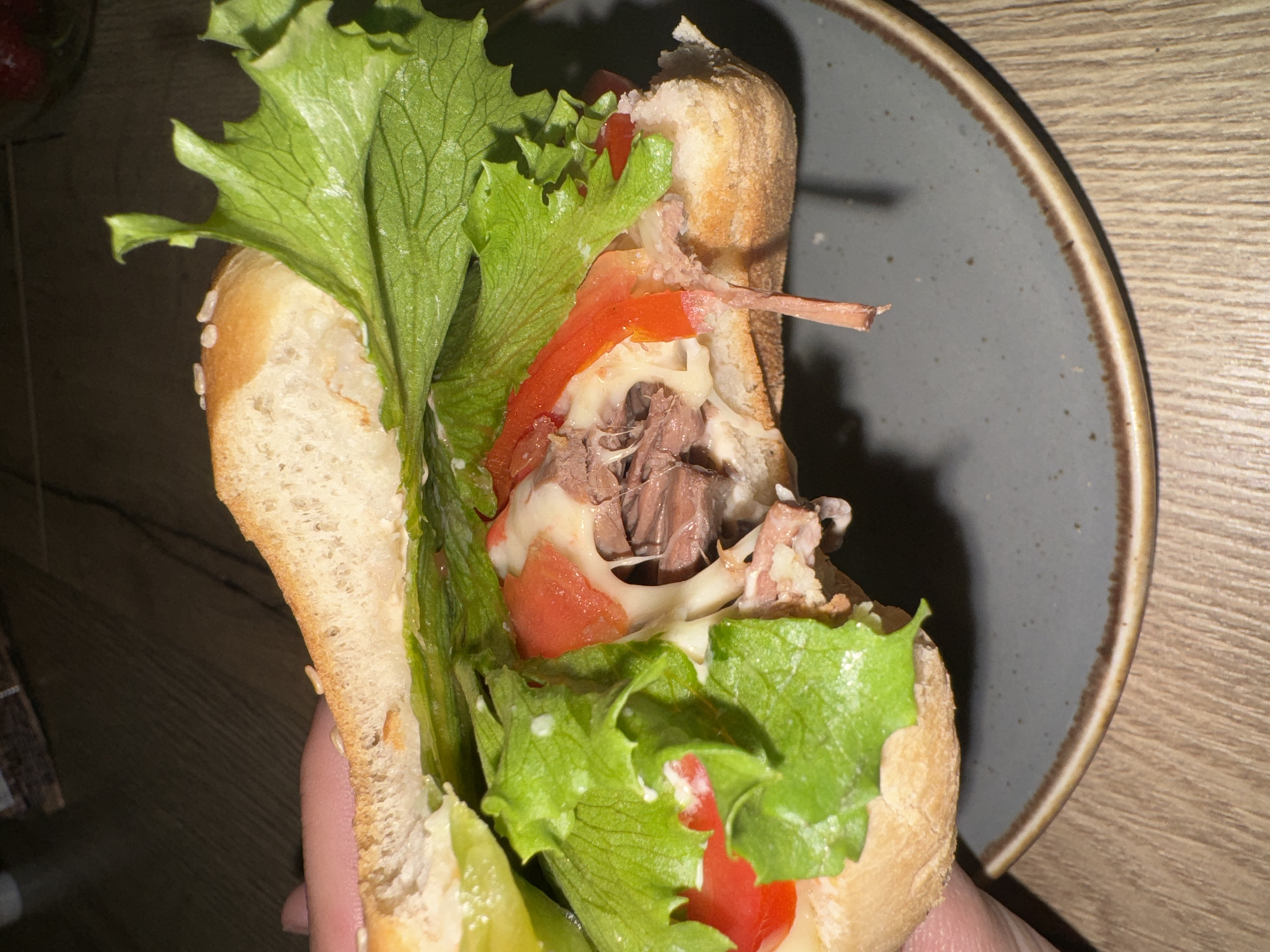 A close-up, slightly overhead shot captures a large, generously filled sandwich being held by a person's hand. The sandwich features a substantial, light-colored bun, possibly with sesame seeds visible on its crust. Inside, there's a vibrant assembly of ingredients: fresh, crisp green lettuce leaves, bright red tomato slices, and a considerable amount of shredded or pulled dark meat, likely beef. A creamy white sauce or melted cheese is generously draped over the meat and tomatoes, adding to the richness of the filling.
The hand, with visible fingers, supports the sandwich from underneath. The sandwich is positioned over a dark grey ceramic plate with a contrasting brown rim, which in turn rests on a light-colored wooden surface, presumably a table. The background is dimly lit and out of focus, revealing a blurry suggestion of a glass or jar containing red items, possibly strawberries, in the upper left corner. The scene depicts an indoor setting, likely a home or casual dining spot, with no visible text or explicit geographical indicators within the image.