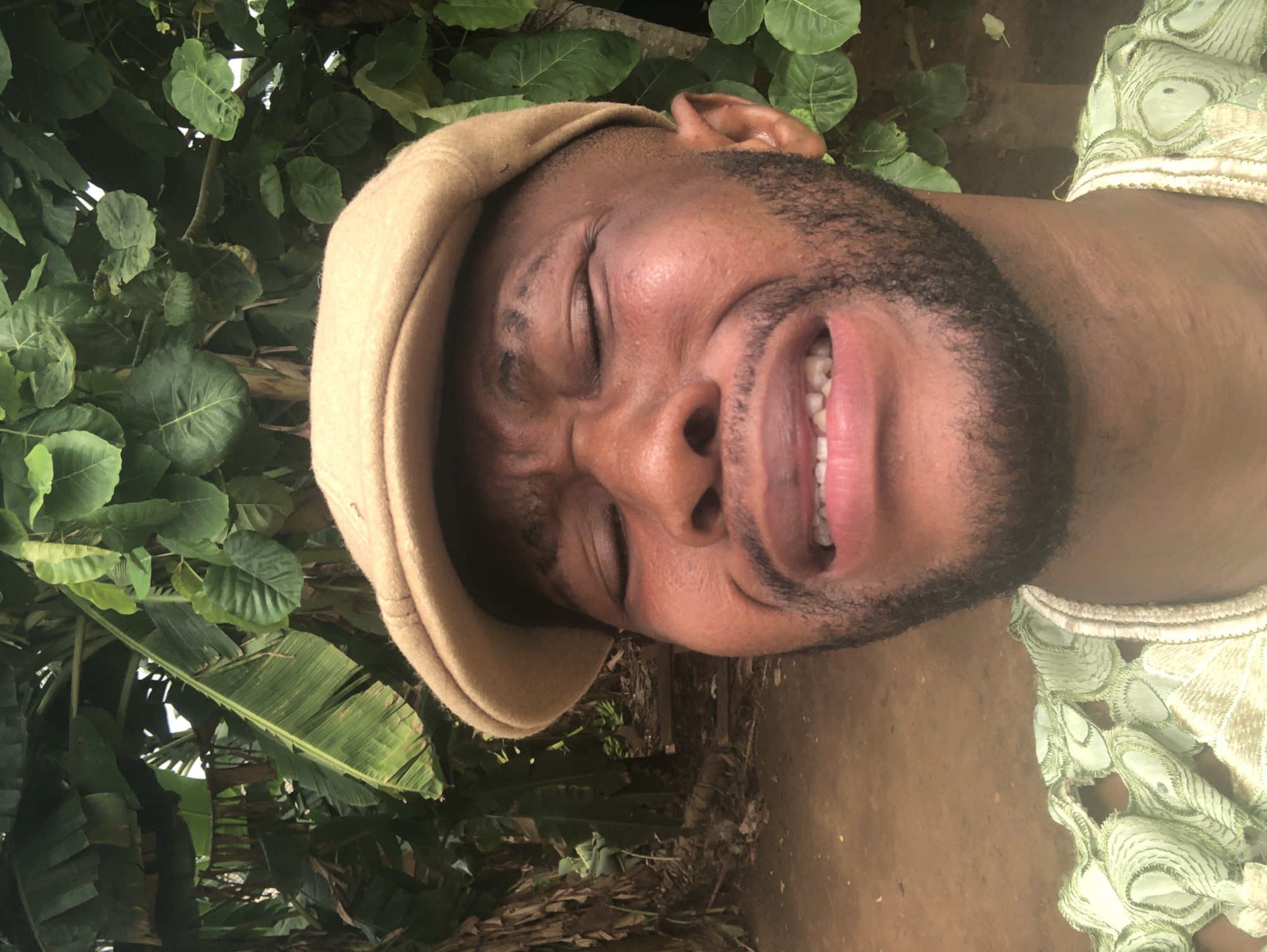 The image features a close-up portrait of a man with dark skin, positioned upright, against a vibrant green natural background. He is captured mid-expression, with his eyes tightly closed or squinting and his mouth open wide, revealing his teeth in what appears to be a strong laugh or a highly animated reaction. He sports a beard or stubble and is wearing a light tan or beige flat cap on his head. His attire includes a light green garment with intricate white or gold embroidered patterns visible on his shoulder and upper chest. The background is lush with various green plants, including large broad leaves, possibly banana leaves, suggesting a tropical or dense natural outdoor setting. Patches of brown earth are also visible behind him. The lighting is bright, indicating it is daytime. There is no visible text in the image.
