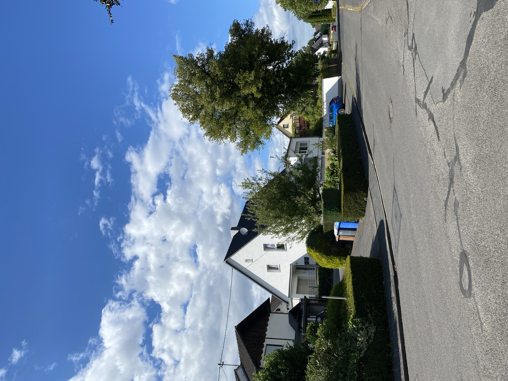 The image depicts a street scene in the city of Flammersfeld, Germany, on a clear day with scattered white clouds against a bright blue sky. The foreground shows an asphalt road with some visible cracks and a manhole cover. On the right side of the road, a gentle slope leads upwards towards a residential area.
The middle ground features several houses with pitched roofs and well-maintained gardens. One prominent white house with a black roof is situated centrally, with a driveway and garage. To its left, another house with a darker roof and wooden accents is visible. In front of the houses, meticulously trimmed green hedges line the property boundaries and parts of the sidewalk. Two residential trash bins, one blue and one brown, are placed next to the hedges. A bright blue car is parked in the driveway of one of the houses. A large, mature tree with dense green foliage dominates the right side of the middle ground, casting a shadow onto the road.
In the background, more houses are visible, stretching along the street as it curves away. Two cars, one red and one dark, are seen further down the road. The environment appears to be a quiet suburban neighborhood, characterized by neat landscaping and residential architecture. The overall impression is one of a peaceful, sunny day in a German town.