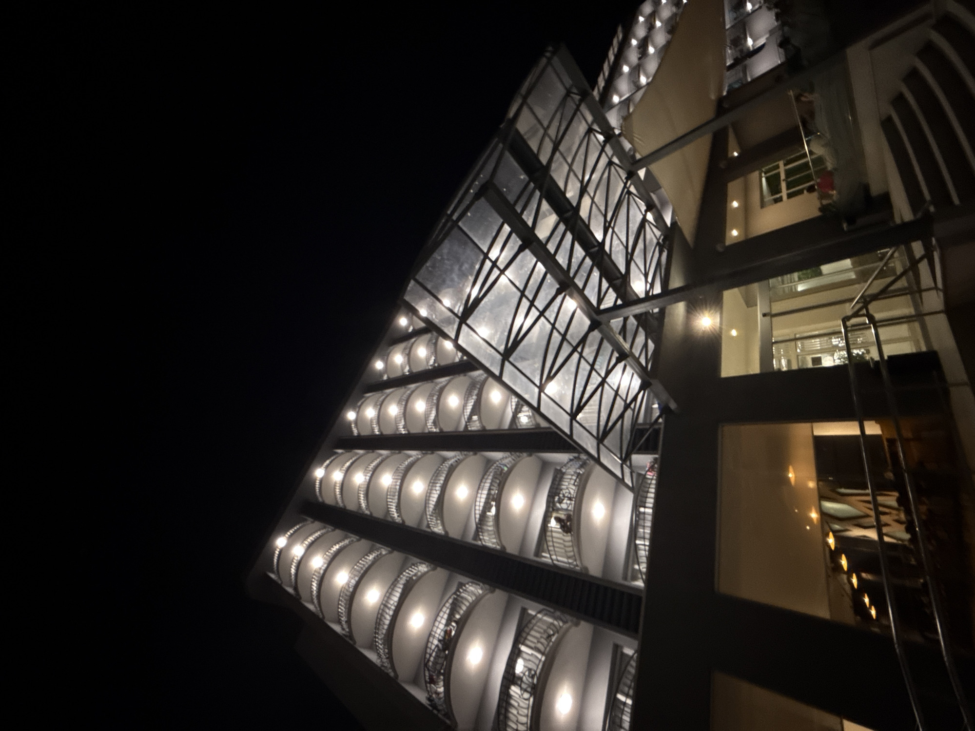 The image shows a modern hotel building at night, likely located in Varna, Bulgaria. The hotel features multiple floors with curved balconies, each illuminated by a central light fixture. A prominent glass and metal canopy extends over the entrance area, with its structure visible through the transparent panels. Inside the building, visible through large windows and glass doors, appear to be the lobby and perhaps a restaurant or seating area, with warm lighting and glimpses of people. The overall impression is one of contemporary architecture and nighttime ambiance.
