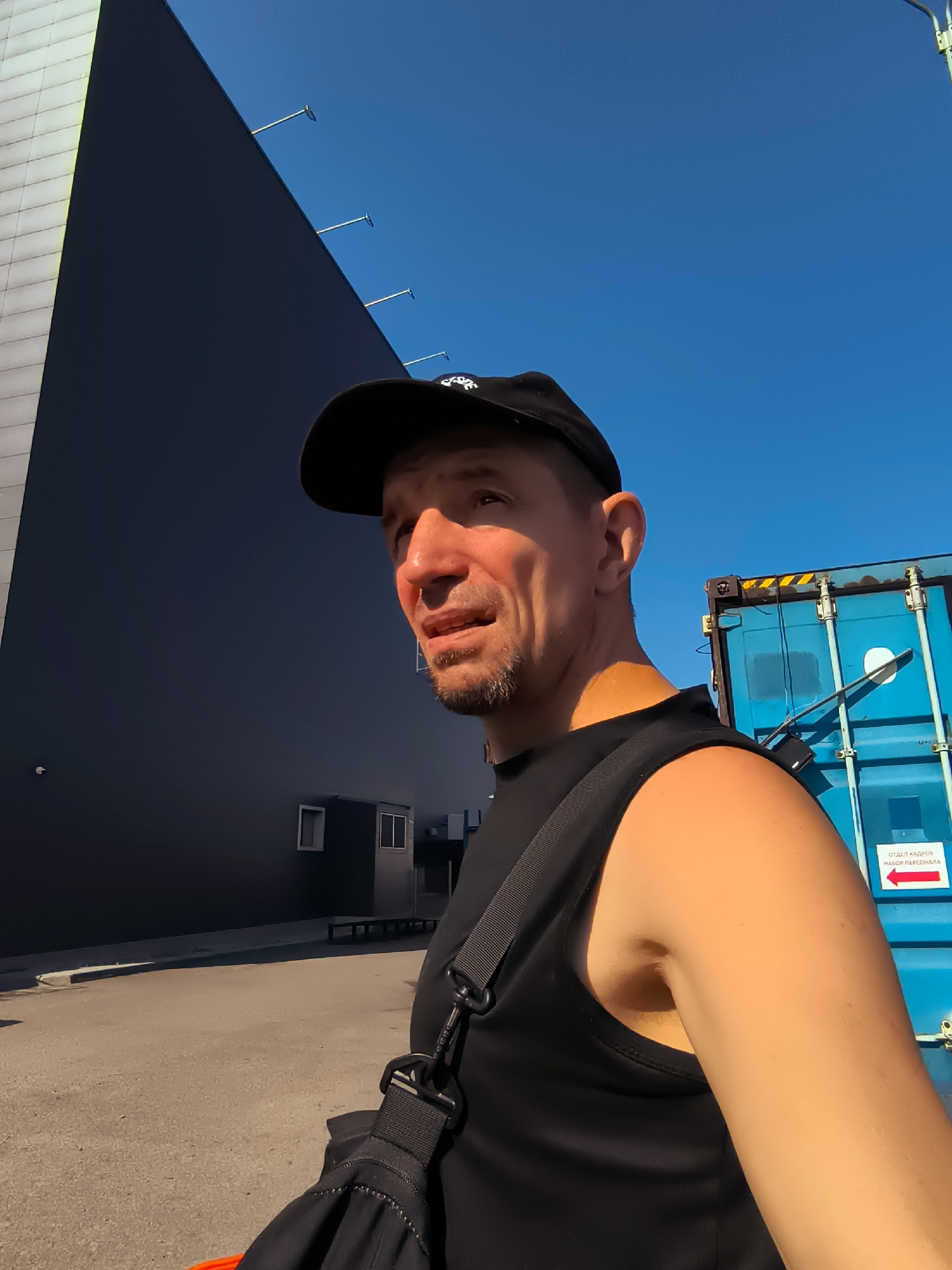 A man wearing a black baseball cap and a black sleeveless shirt is standing outdoors. He has a light beard and is looking to his right. Behind him, to the left, is a large dark building with a white section on the far left. To his right is a blue shipping container with some writing and a red arrow on a white sticker. The sky is clear and blue. The scene appears to be an industrial or commercial area. There is no discernible information to place this image in Qarağandı city administration, Kazakhstan.