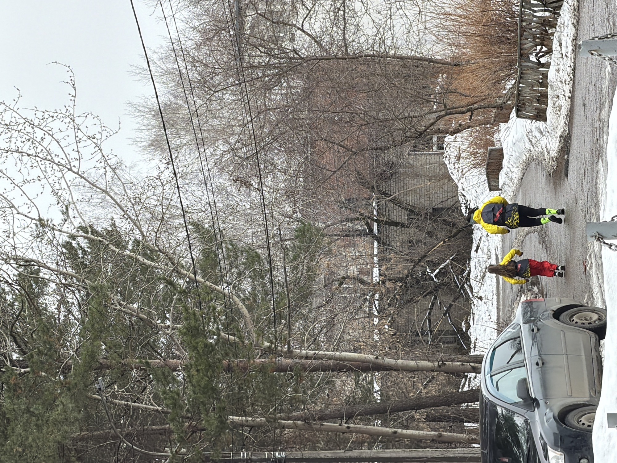 The image depicts a street scene in what appears to be a residential area in Chaikovsky, Russia during the late winter or early spring season. Two individuals, both wearing bright clothing (yellow jackets with red pants) and what look like roller skis, are roller skiing down a snow-lined street. A dark-colored car is parked at the edge of the street. A brick building is visible in the background, and bare trees and power lines are overhead. Patches of snow are visible alongside the road and on the adjacent building.