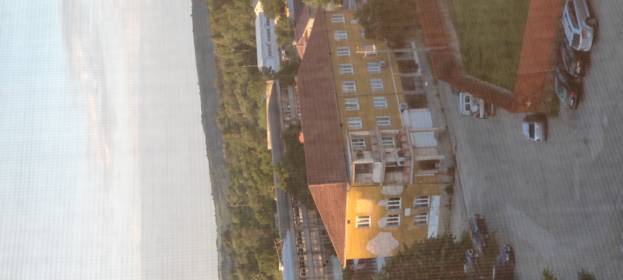 The image captures a partially obstructed aerial view of a cityscape. A large, yellow building with a red roof stands prominently, showing signs of construction or renovation on some of its floors. Adjacent to the building, there's a green area and a parking lot occupied by multiple cars. The background reveals a dense forest and other buildings in the distance. The overall setting appears to be a residential or suburban area.