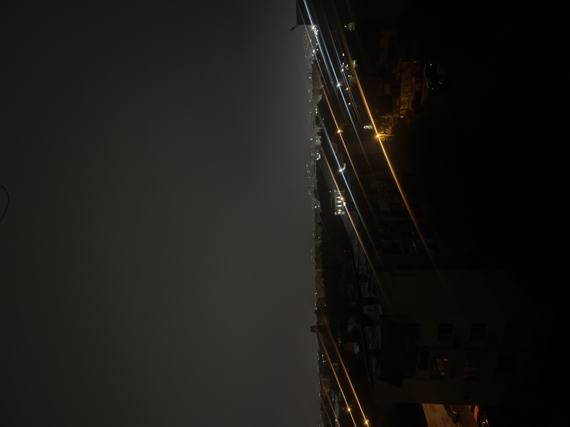 The image depicts a nighttime cityscape in Sofia, Bulgaria. The view is oriented vertically, with a dark, indistinct foreground and a lighter horizon. Streaks of light indicate streetlights and vehicle headlights extending into the distance. Buildings are visible as dark shapes along the right side. A substantial portion of the left side is a dark, clouded sky. There are no discernible people or specific actions depicted.