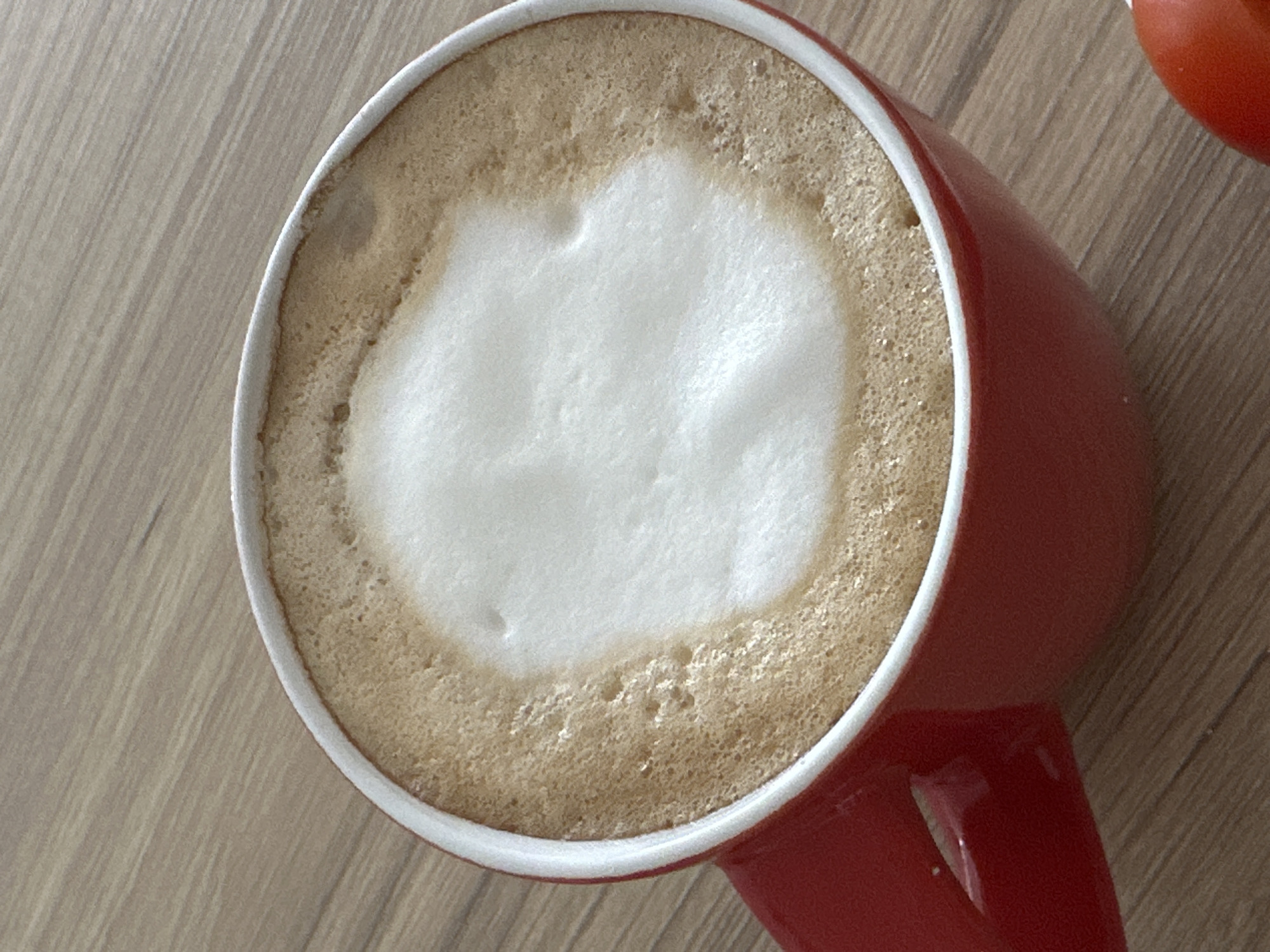 A top-down, close-up shot captures a red ceramic mug filled with a cappuccino. The cappuccino boasts a thick layer of foamed milk, with the central foam being noticeably whiter and denser than the outer ring, which is tinted with coffee. The mug sits on a wooden surface, possibly a table. A red object, partially visible in the upper right corner, is unidentified. The location in the city of Budva, Montenegro is not visible within the frame of the image.