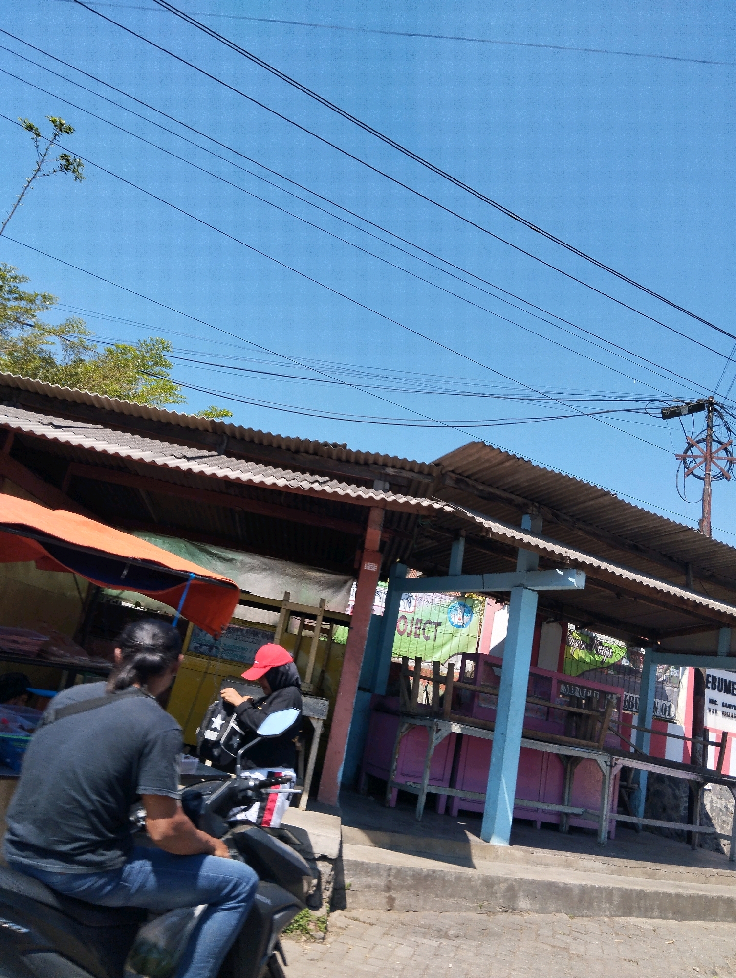 The image depicts a street scene in Krajansari, Indonesia, under a clear blue sky crisscrossed with numerous overhead power lines. In the foreground, a person wearing a black t-shirt and blue jeans is seated on a black motorcycle, their back to the viewer, with long dark hair. Beside them, a person wearing a red cap and dark clothing is also on a motorcycle, adjusting a black backpack with a white star emblem. Behind them, a corrugated metal roof structure, possibly a market stall or shelter, extends across the frame. The structure has a weathered appearance, with exposed wooden beams and metal supports. A bright orange awning is visible on the left side of the structure. In the background, banners and signs are partially visible, including one with the word "PROJECT" and another with text that appears to be in Indonesian. The ground in front of the structure is a paved walkway with a concrete curb. The overall impression is of a bustling, everyday scene on a bright, sunny day.