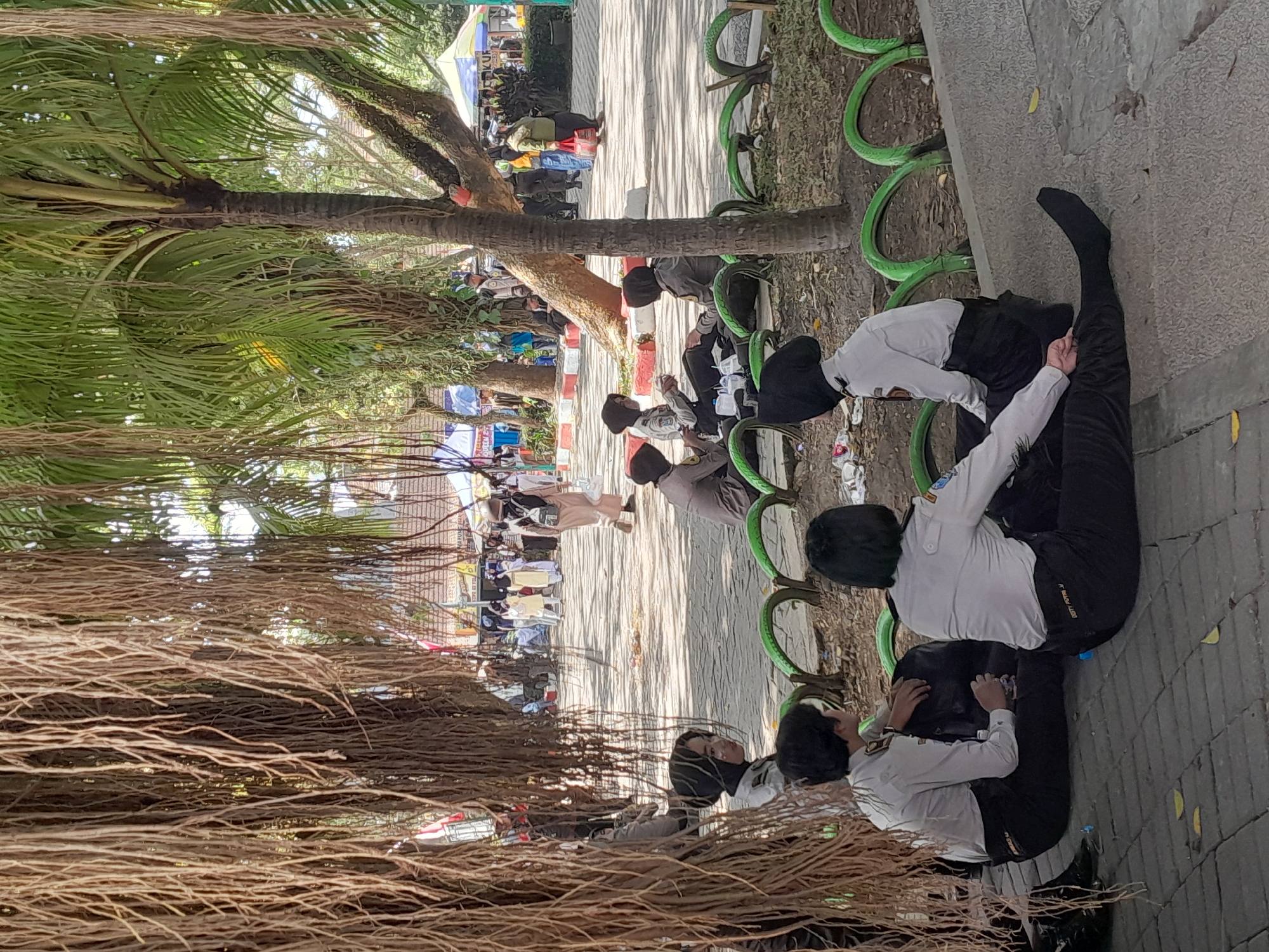 In Genteng Kulon, Indonesia, a group of young people, likely students in uniform, are gathered outdoors. The scene is set in a park-like area with trees, including one with visible aerial roots hanging down in the foreground, and a palm tree. The individuals are seated on a paved surface, some on the ground and others on a slightly raised ledge. They are dressed in light-colored long-sleeved shirts and dark pants, with some wearing headscarves. Some are interacting with each other, while others appear to be resting or observing. In the background, there are more people walking, tents, and structures, suggesting an event or gathering is taking place. The ground is a mix of paving stones and dirt, with some green, semi-circular barriers visible in the middle ground.