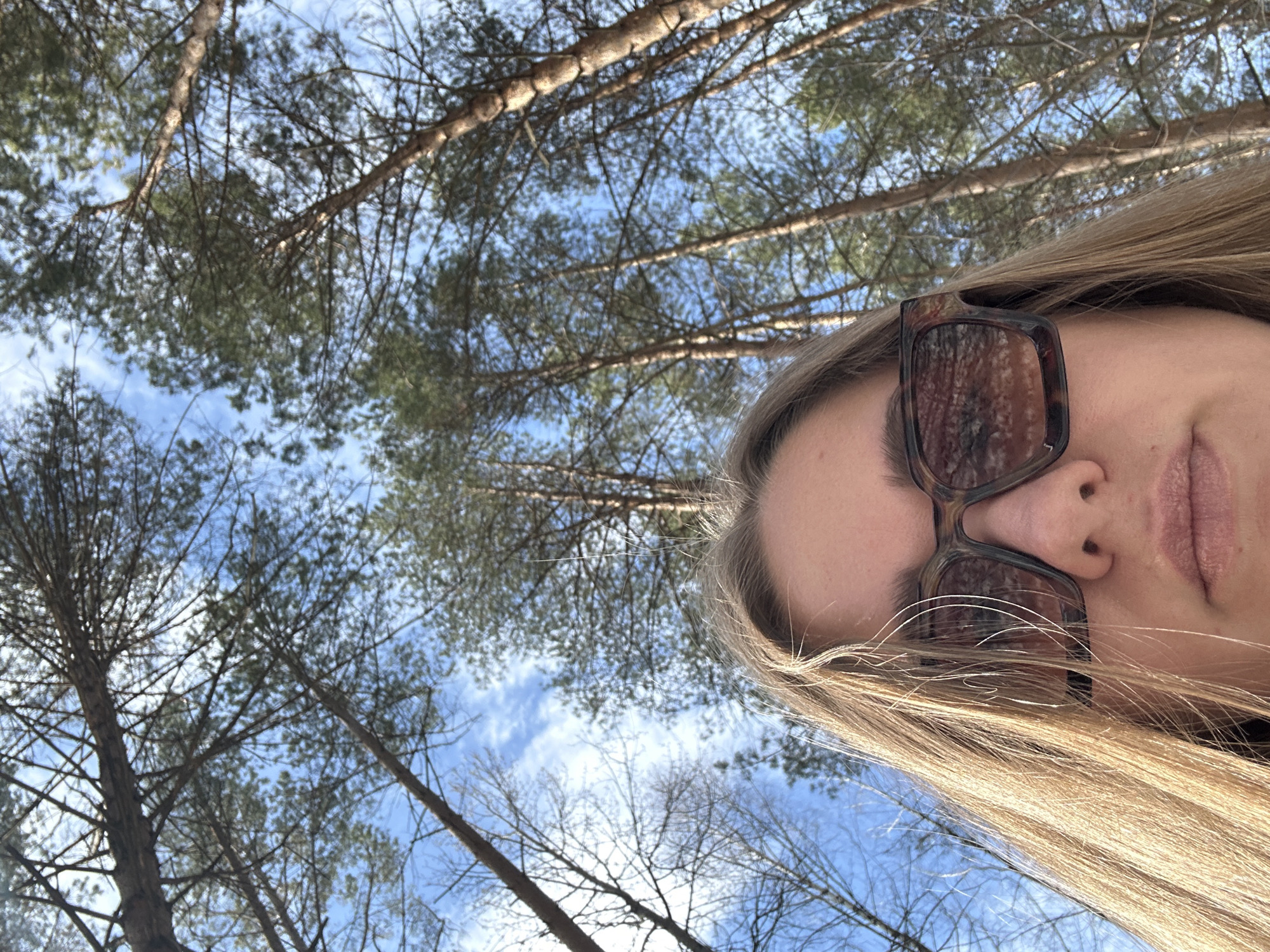 The image is a low-angle selfie of an individual, likely female, set in a forest environment under a clear blue sky with scattered white clouds. The lower portion of the subject's face, including the nose, mouth, and chin, is visible in the foreground, with the eyes obscured by dark-framed, possibly tortoiseshell-patterned, sunglasses. Light-colored hair frames the face. The background consists of numerous tall trees, predominantly conifers with green needles, ascending towards the sky due to the low camera angle. Some bare deciduous tree branches are also visible on the left. The scene suggests a wooded area or park, located in the city of Chaikovsky, Russia, during a season such as winter or early spring.