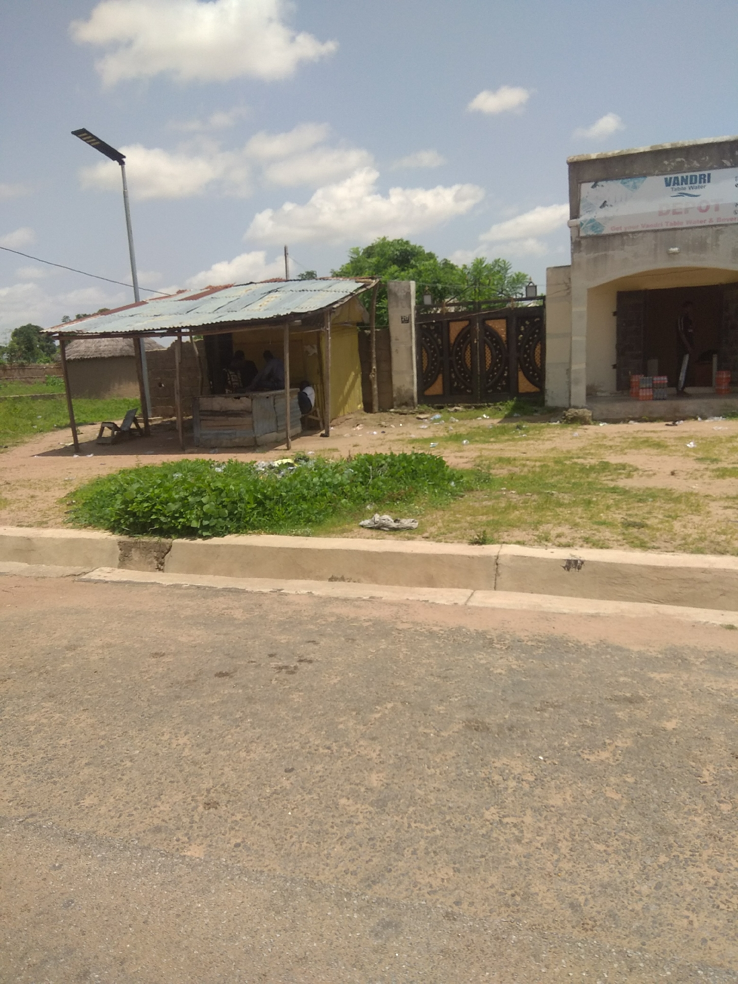 This is a daytime, eye-level, outdoor photograph taken in Jalingo, Nigeria. The scene depicts a street-level view with a road in the foreground, a curb, and a dusty area with vegetation. To the left, a small, rustic structure with a corrugated metal roof and wooden supports serves as a shop or stall. Several people are gathered inside this structure. In front of the shop, there is a chair. To the right of the shop, a larger, more permanent building with a tan facade and an arched entrance is visible. A large, ornate metal gate with intricate patterns stands between the shop and the building. The sky above is blue with scattered white clouds, suggesting fair weather. A tall, modern street lamp is also present in the scene. There is some litter scattered on the ground, particularly near the gate and in front of the shop. The building on the right has a sign that reads "VANDRI TABLE WATER DEPOT". The overall impression is of a commercial area in a developing locale, possibly a market or a roadside business district.