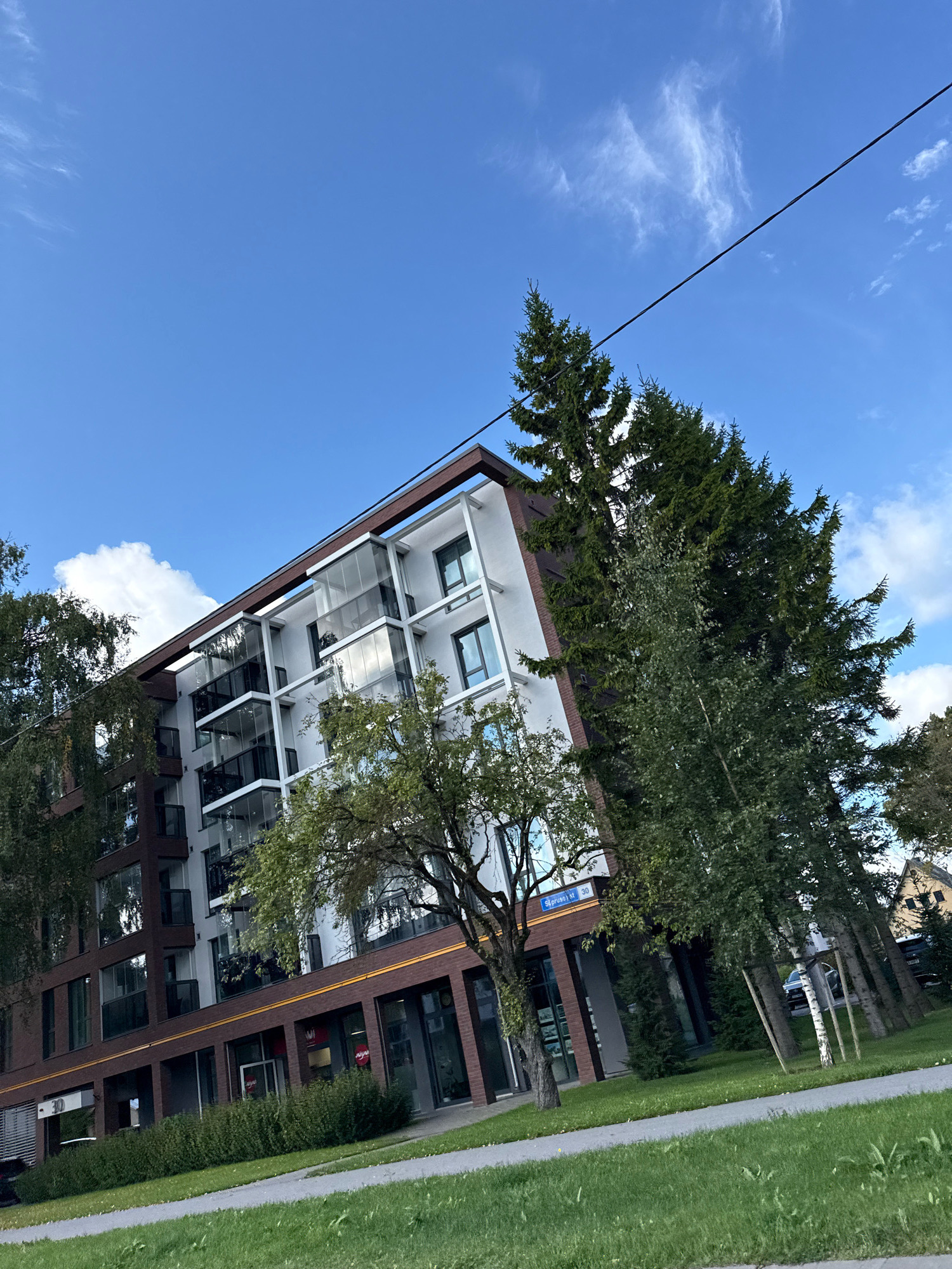This image shows a modern apartment building with a dark brown brick facade on the lower floors and white upper floors with glass balconies. The building is surrounded by lush green trees, including tall coniferous trees and deciduous trees with green foliage. A paved walkway and a grassy area are in the foreground. The sky is blue with scattered white clouds, and a power line stretches diagonally across the upper portion of the image. A street sign reading "Struves iela 30" is visible on the building, indicating the location is Struves Street 30 in Tallinn, Estonia. There are no people visible in the image. The overall scene depicts a well-maintained residential area on a clear day.