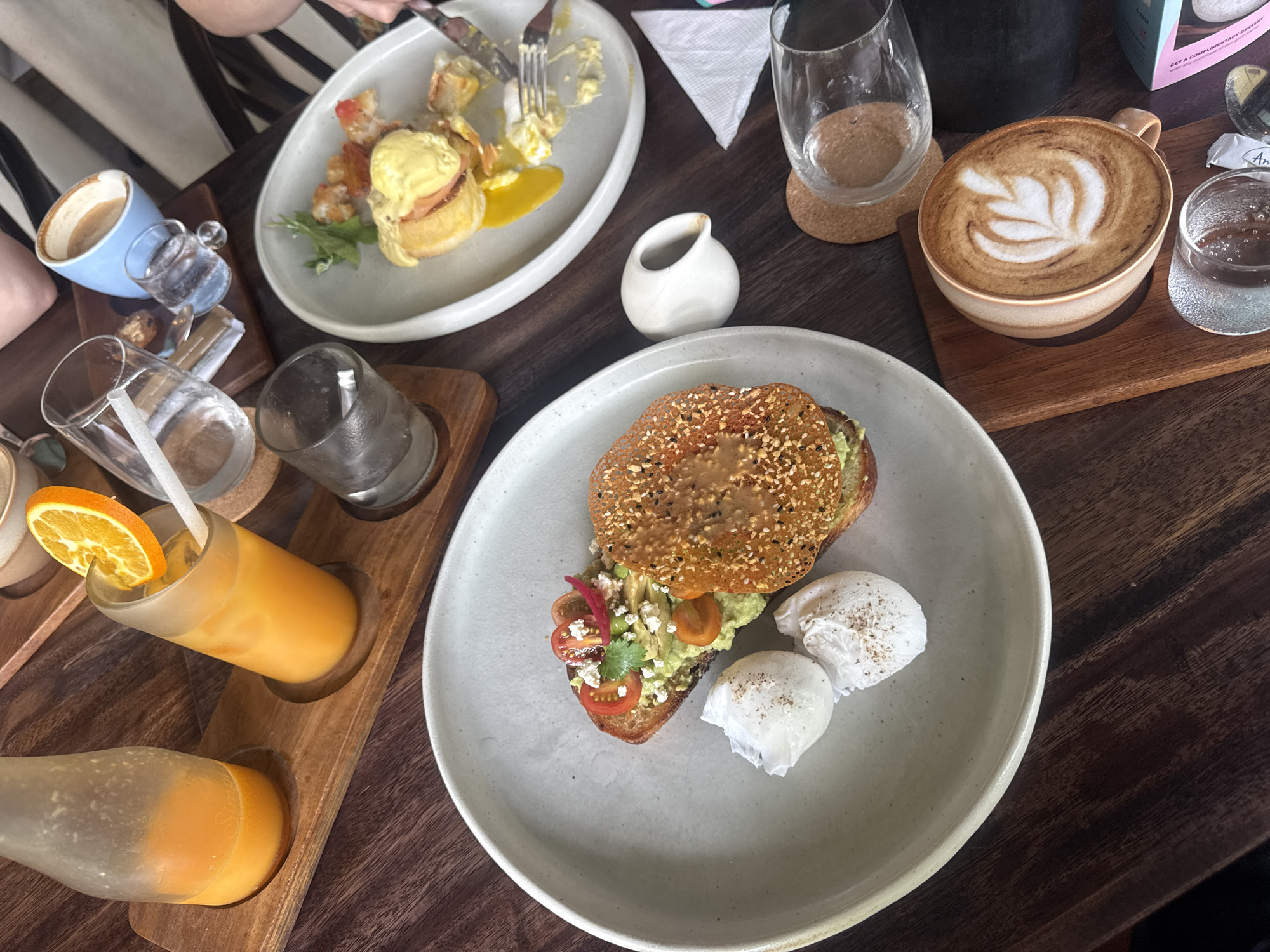 A top-down shot captures a vibrant breakfast or brunch spread for multiple diners at a cafe in Ubud District, Indonesia. The dark wooden table is laden with various dishes and drinks, suggesting a relaxed and enjoyable meal.
In the foreground, a large, light grey ceramic plate holds a healthy avocado toast. This features a thick slice of seeded toast topped with creamy avocado, colorful cherry tomatoes, crumbled white cheese, and a sprig of cilantro. Beside it are two perfectly poached eggs, lightly seasoned with pepper.
Towards the top left, another light grey plate showcases a classic Eggs Benedict, with golden hollandaise sauce over what appears to be ham or bacon on a base, accompanied by scrambled eggs or hash. A person is seen actively eating from this plate with a fork and knife.
Beverages abound: a latte or cappuccino with intricate latte art sits in a beige cup on a wooden coaster, next to a small glass of water. A blue ceramic mug with a beige rim contains another drink. To the left, a wooden tray holds two glasses or bottles of orange juice, one garnished with a fresh orange slice and a straw. Several empty or partially filled water glasses are also visible, along with a small white creamer.
Notable details include a white paper napkin, small wrapped sugar packets (one with visible text "Anda"), and a partially visible light blue box with text "GET a complimentary dessert", "sweet", and "ANY", hinting at a promotion or branding. The overall scene evokes a pleasant and satisfying dining experience, likely during the morning or early afternoon.
