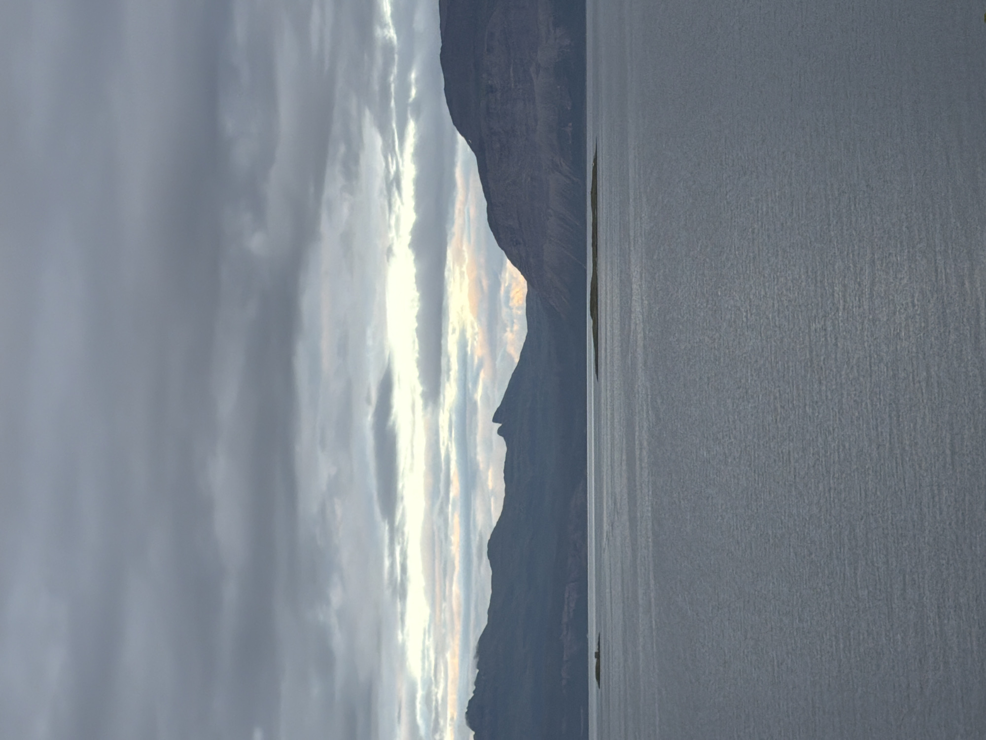 The image shows a wide expanse of calm water, likely a fjord or bay, with a dark, mountainous landscape dominating the background. The mountains are rugged and appear to be covered in a dark, muted color, possibly due to vegetation or rock type. The sky is overcast with a thick layer of gray clouds, though there are breaks in the clouds near the horizon where hints of soft yellow and pink light can be seen, suggesting either dawn or dusk. There are a few small, low-lying islands or rocky outcrops visible in the water, one of which is closer to the viewer on the left, and another larger one further in the distance. The water itself has a slightly textured surface, indicating very gentle ripples. The overall atmosphere is serene and somewhat somber, characteristic of a moody Nordic landscape. This scene is typical of the natural beauty found in and around Harstad, Norway, which is surrounded by fjords and mountains.