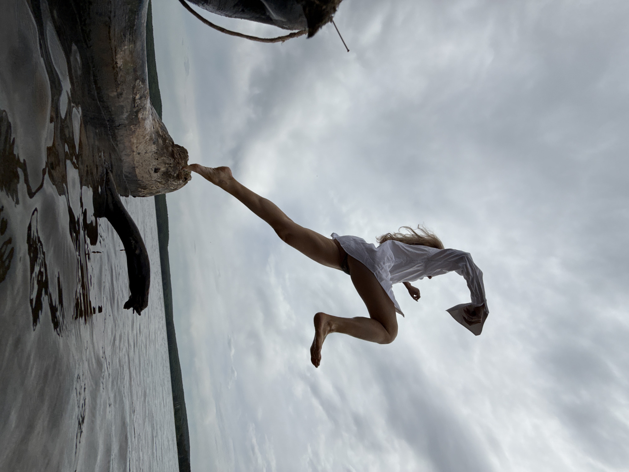 A low-angle, color photograph depicts a woman in mid-air, wearing a white, long-sleeved shirt and dark-colored undergarments, leaping away from a section of a large, weathered tree trunk extending over water. The woman's right foot is in contact with the tree, while her body is angled towards the sky. The background consists of an overcast sky and the body of water. The location appears to be a natural setting, possibly near a lake or river, near Chaikovsky, Russia.