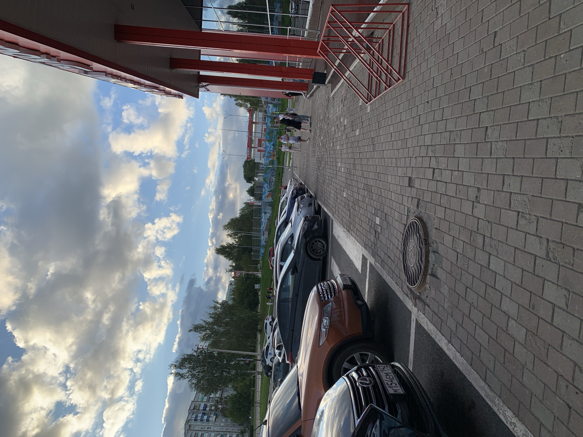 A wide-angle, eye-level outdoor photograph captures a parking lot scene in Prokopyevsk, Russia. The scene is dominated by a row of parked cars and a sidewalk paved with interlocking bricks. In the background, a modern building with red columns and large glass windows is visible, suggesting a commercial or public establishment. The sky above is filled with dramatic clouds, indicating either sunrise or sunset. Several people are seen walking on the sidewalk near the building. The parking lot itself is marked with white lines, and a manhole cover is present on the ground. The overall atmosphere is one of urban activity under a dynamic sky.
