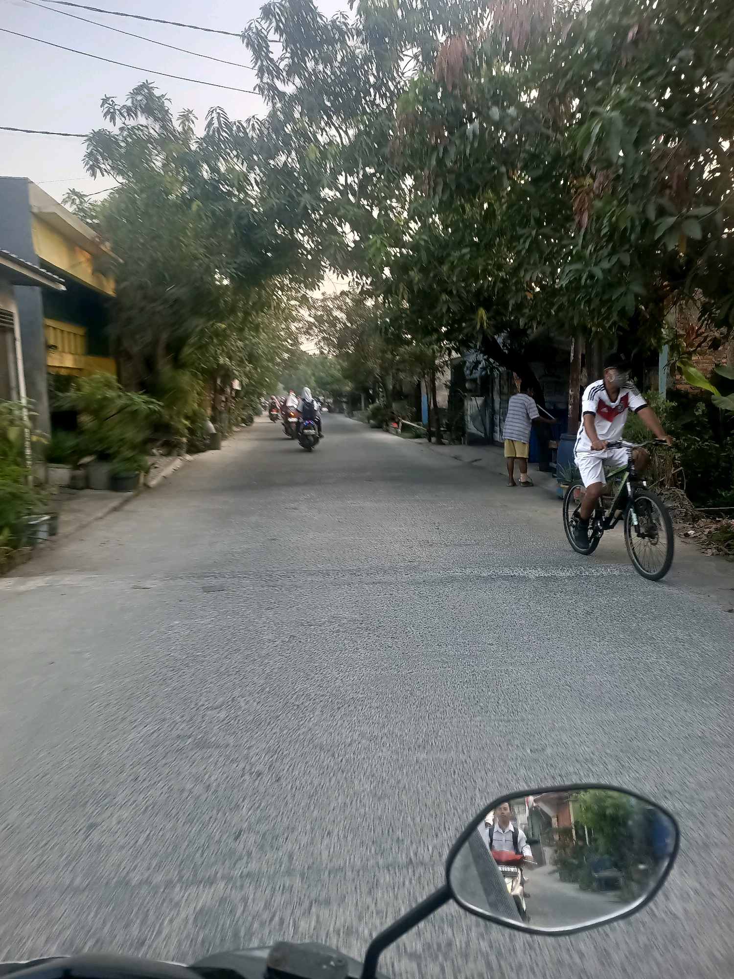 A man wearing a white shirt with red and black trim, white shorts, and a face mask rides a bicycle down a paved road in Wanajaya, Indonesia. The bicycle is black and green. Another person, wearing a striped t-shirt and yellow shorts, stands near a building on the side of the road. Several motorcycles are visible further down the road. Trees line both sides of the road, with houses and other structures interspersed among them. The image appears to have been taken from the perspective of someone riding a motorcycle, as a rearview mirror is visible in the foreground, reflecting the rider. The sky is overcast, and the light suggests it is either early morning or late afternoon.