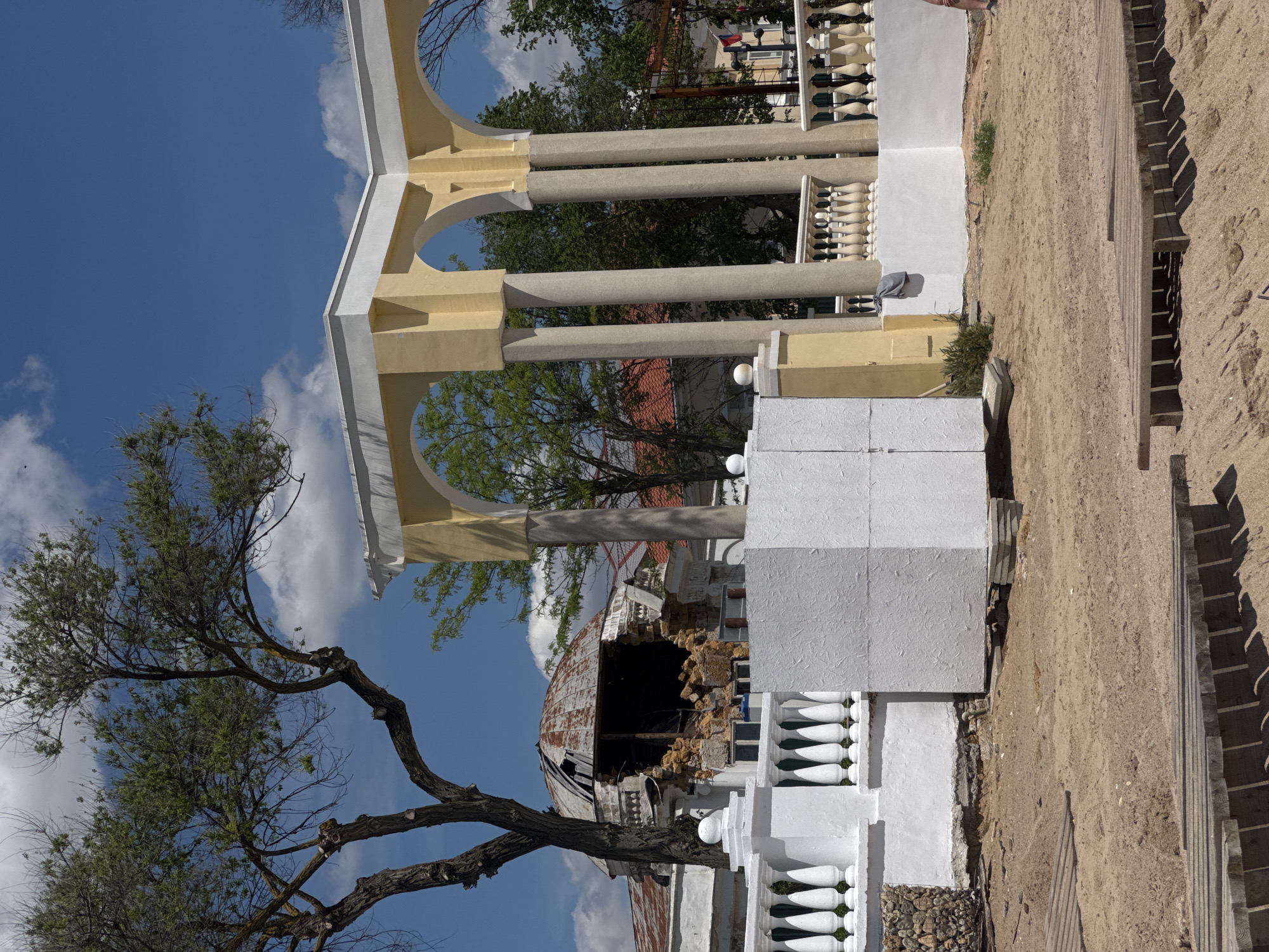 The image shows damage from an apparent explosion or other destructive event at what appears to be a seaside structure in Yevpatoriya, Ukraine. The scene includes a large white and yellow colonnade with arches, suggesting a decorative or recreational building, potentially a gazebo or pavilion. To the side, there is a severely damaged structure with a collapsed dome, now exposing crumbled brickwork and debris. A tree with bare branches partially obscures the foreground. White painted railings are visible next to the damaged structure. Fragments of debris and building materials are scattered on the ground. The background shows glimpses of other buildings and trees under a partly cloudy sky.