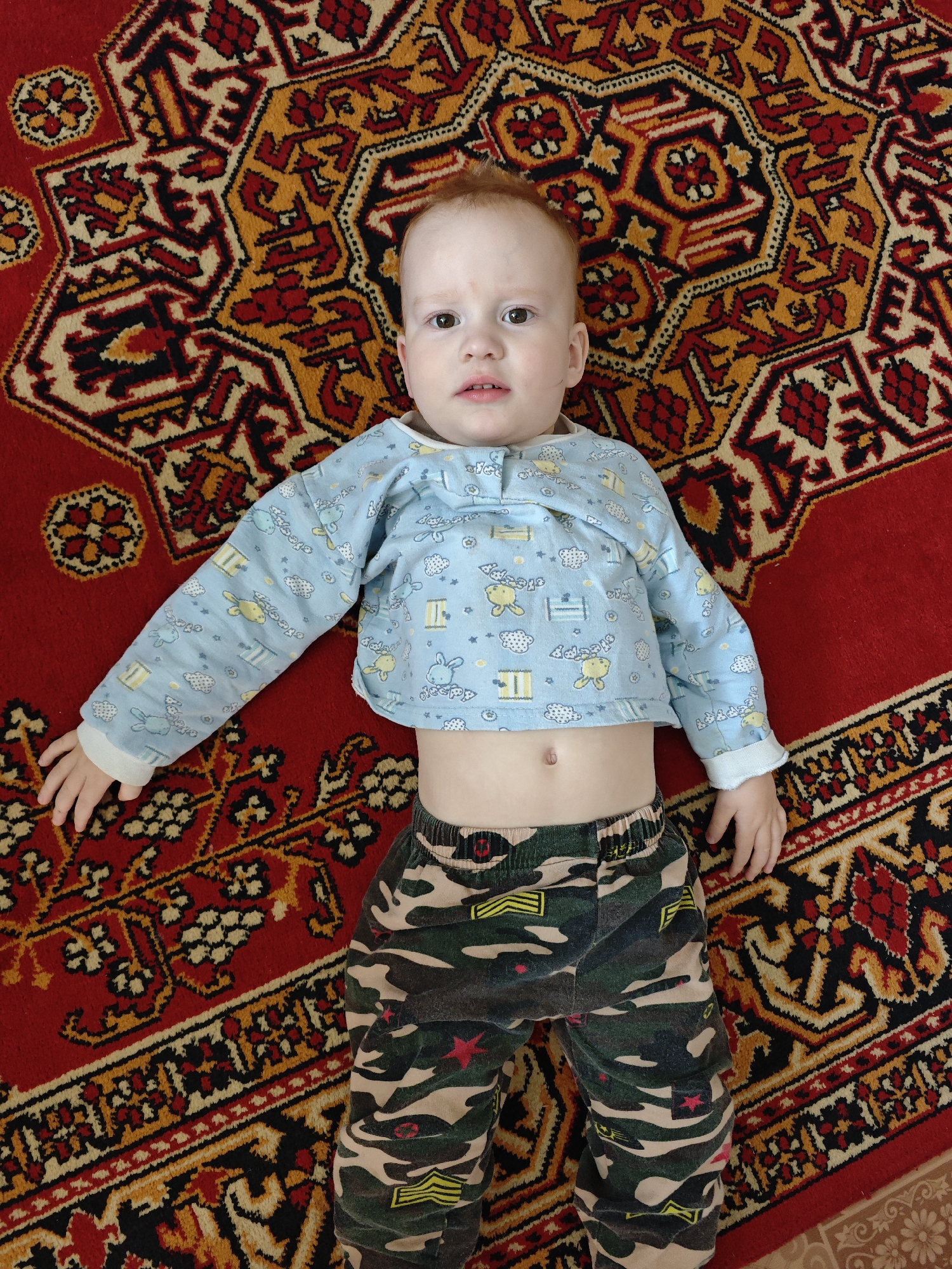 A baby with light red hair is lying on a carpet with an intricate, colorful pattern. The baby is wearing a light blue top with cartoon illustrations and camouflage patterned pants. The location appears to be inside a building, based on the presence of the carpet. The city of Фоки, Russia, is not apparent from visual information.