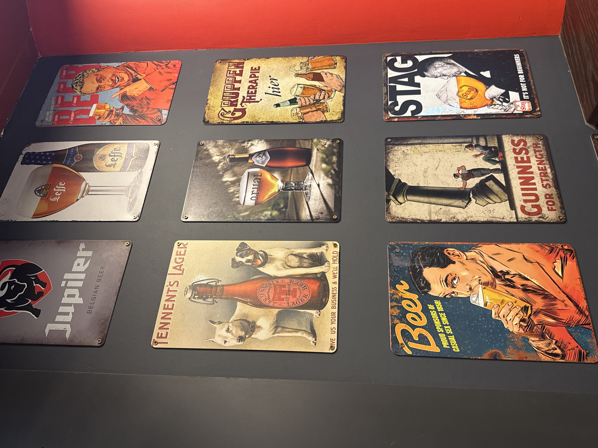 Nine vintage-style metal advertising signs for various beer brands are displayed on a dark grey wall, with a red trim visible along the top edge of the wall, suggesting an indoor setting in Saint Petersburg, Russia, likely a pub or home bar. The signs are arranged in three rows and three columns, creating a collage.
The signs depict various subjects and themes:
* **Top left:** A grinning man points to a glass of beer, with text "BEER" and days of the week listed.
* **Top center:** Hands holding various beer glasses and bottles, with text "GRUPPEN THERAPIE hier" (Group Therapy here).
* **Top right:** A black-and-white graphic of a man drinking from a glass of Stag beer, with text "STAG IT'S NOT FOR BEGINNERS".
* **Middle left:** A Leffe beer bottle and a chalice glass filled with beer, marked "Leffe ANNO 1240".
* **Middle center:** An Orval beer bottle and a chalice glass, with "Orval" visible.
* **Middle right:** Two men attempting to move a large, muscular arm holding a Guinness barrel, with text "GUINNESS FOR STRENGTH".
* **Bottom left:** A Jupiler beer sign featuring a bull's head logo and "Jupiler BELGIAN BEER".
* **Bottom center:** Two dogs flanking a Tennent's Lager bottle, with "TENNENT'S LAGER" and "GIVE US YOUR BUSINESS & WE'LL HOLD" (partially visible).
* **Bottom right:** A comic-book style illustration of a man drinking a beer, with text "Beer PROUD SPONSORS OF CASUAL SEX SINCE 1858!".
All signs exhibit a distressed, aged aesthetic with faux rust and faded elements, enhancing their vintage appearance. They are affixed to the wall with visible screws in each corner.