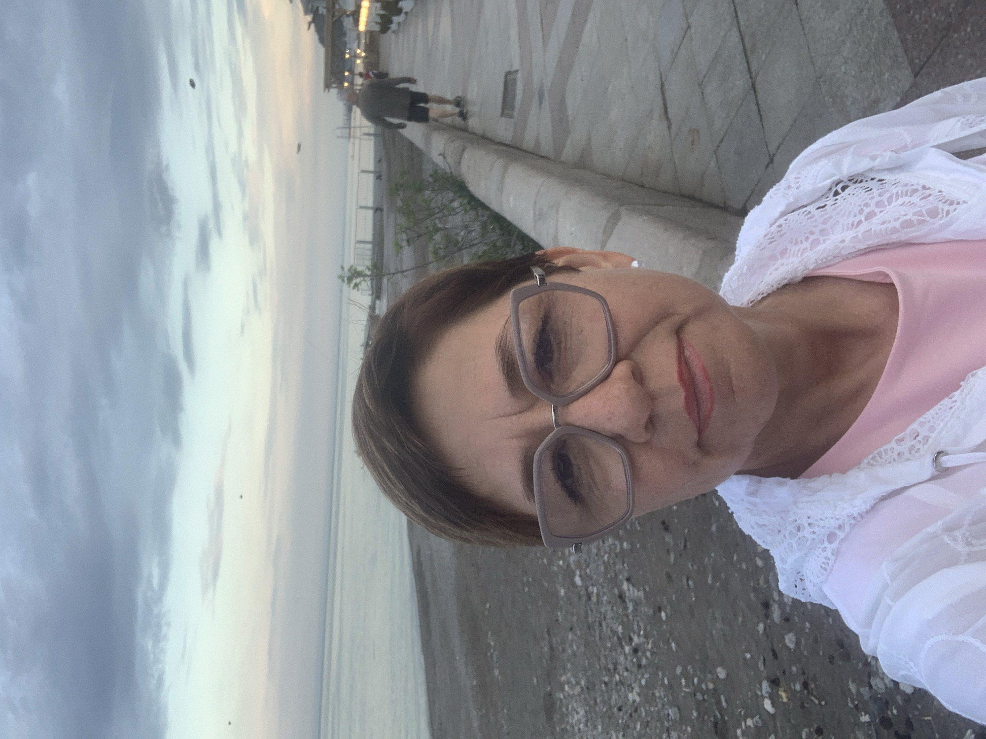 A medium eye-level close-up shows a woman with short brown hair and glasses looking slightly down at the camera. She's wearing a pink shirt and a white lace jacket. The background includes a pebble-strewn beach, the ocean, and a promenade with a person walking in the distance. The sky is overcast with hues of grey and some lighter tones. The location appears to be along a coastal promenade.