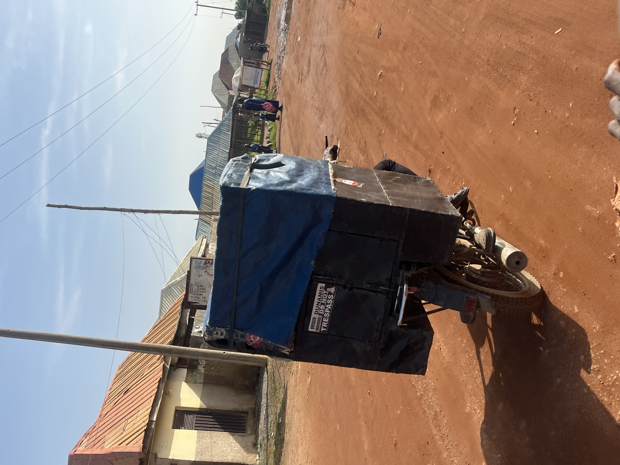 This outdoor image captures a sunny day on a reddish-brown dirt road in what appears to be a semi-urban or rural area, consistent with the context of Abuja, Nigeria. The scene is dominated by a three-wheeled vehicle, likely a commercial tricycle (often called a Keke Napep), parked on the right side of the road. The rear cargo/passenger section of the tricycle is covered with blue and black material.
In the mid-ground, several people are visible walking or standing near structures and a lighter-colored, possibly paved area. Buildings with corrugated iron roofs line the background. Overhead, a clear sky with a few wispy clouds is visible, crisscrossed by several utility lines extending from a wooden pole on the left. The bright sunlight casts distinct shadows, notably the tricycle's shadow stretching across the dirt road, indicating it is likely midday or early afternoon.
Notable text found on the tricycle reads: "ORIGIN: DUNAMIS" and "WARNING: DO NOT TRESPASS A". A sign in the distance also shows partial text, including words like "ATING", "COPY", "LING", "PLASTIC ED", and "TYPESETTING". The overall impression is a candid snapshot of daily life and transportation in the area.