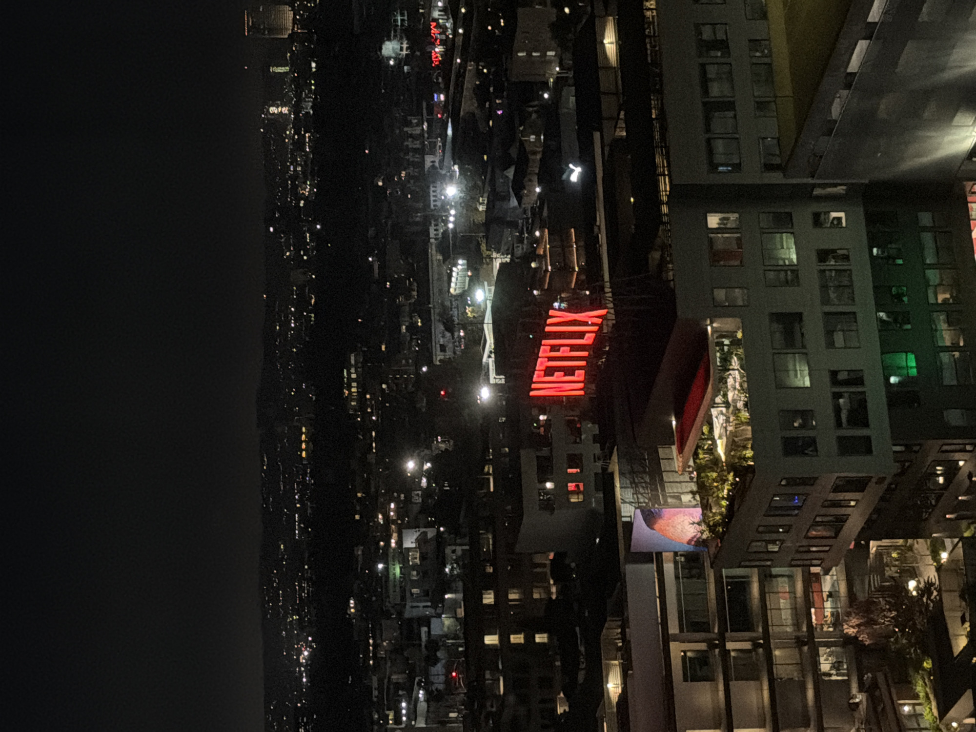 The image shows a nighttime view of the city of Los Angeles, USA, with a focus on the Netflix headquarters. The foreground features a building with a large, illuminated red "NETFLIX" sign on its roof. The sign is composed of individual block letters that emit a bright red light, creating a prominent visual in the otherwise dark cityscape. Surrounding the Netflix building are other urban structures, including multi-story buildings with illuminated windows, indicating occupied spaces. Beyond the immediate vicinity, the city extends into the distance, characterized by a sprawling landscape of lights from other buildings and streetlights, suggesting a dense urban environment. The sky above is dark and clear, typical of a night scene. In the far distance, a few taller buildings are visible, some with lights that stand out against the skyline. The overall impression is an urban nocturnal panorama, with the Netflix sign serving as a central focal point.