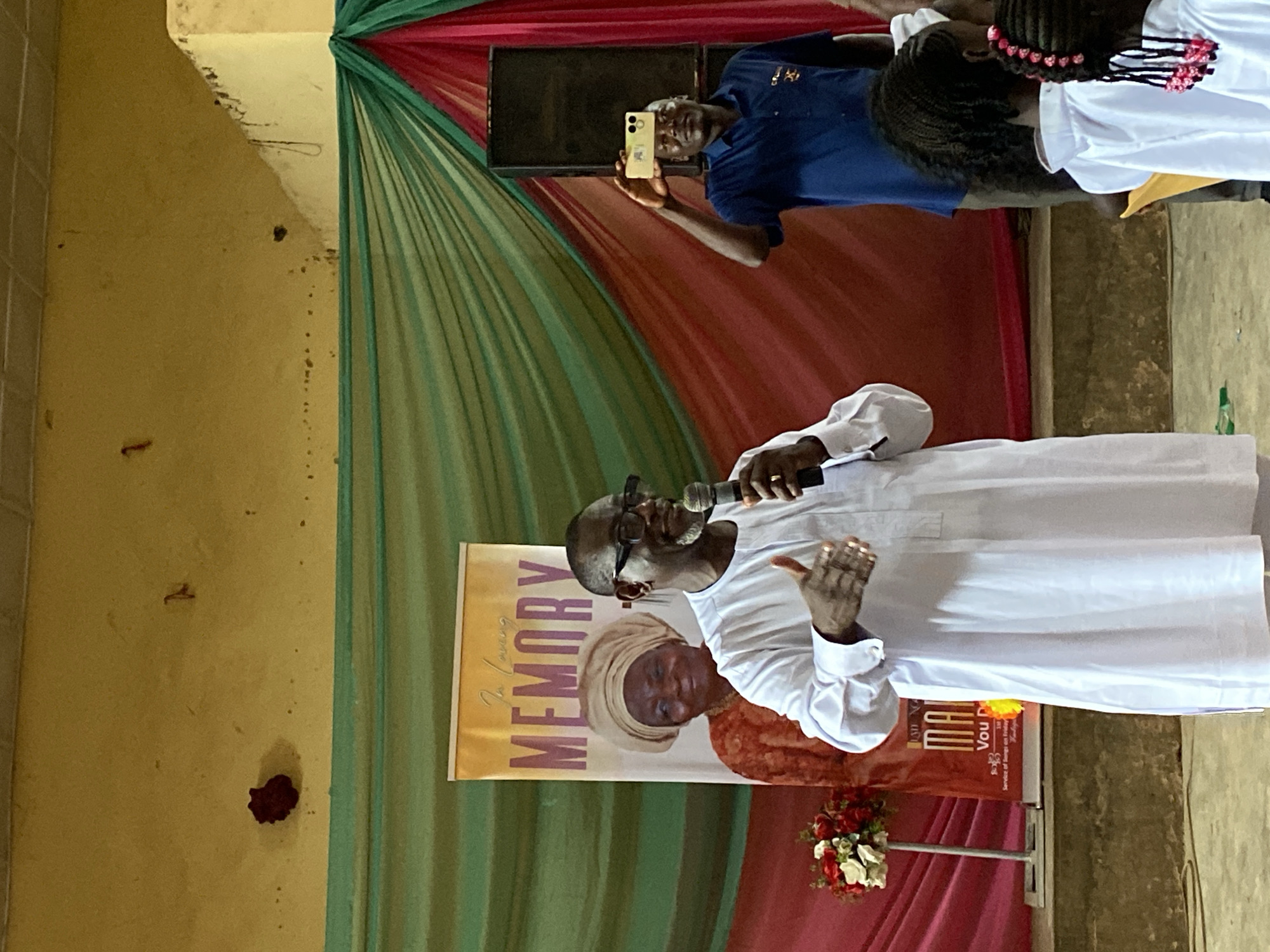 The image depicts an indoor event, likely a memorial service, held in Kuru, Nigeria. The background features draped fabric in shades of green, red, and pink, suggesting a festive or commemorative decoration. A banner with "In Loving MEMORY" is prominently displayed, featuring a portrait of a woman.
In the foreground, a Black man, appearing to be in his 50s or 60s, is speaking into a microphone. He is wearing glasses and a white traditional outfit. His left hand is raised, with his palm facing outward, as if gesturing or addressing the audience. To his right, another Black man, wearing a blue collared shirt, is holding a smartphone up, seemingly recording or taking a picture of the speaker. In the lower right of the frame, the backs of several young Black girls in white outfits are visible, their hair styled in braids. To the left of the speaker, a floral arrangement of red and white flowers on a stand is visible. The overall environment appears to be a hall or community center.