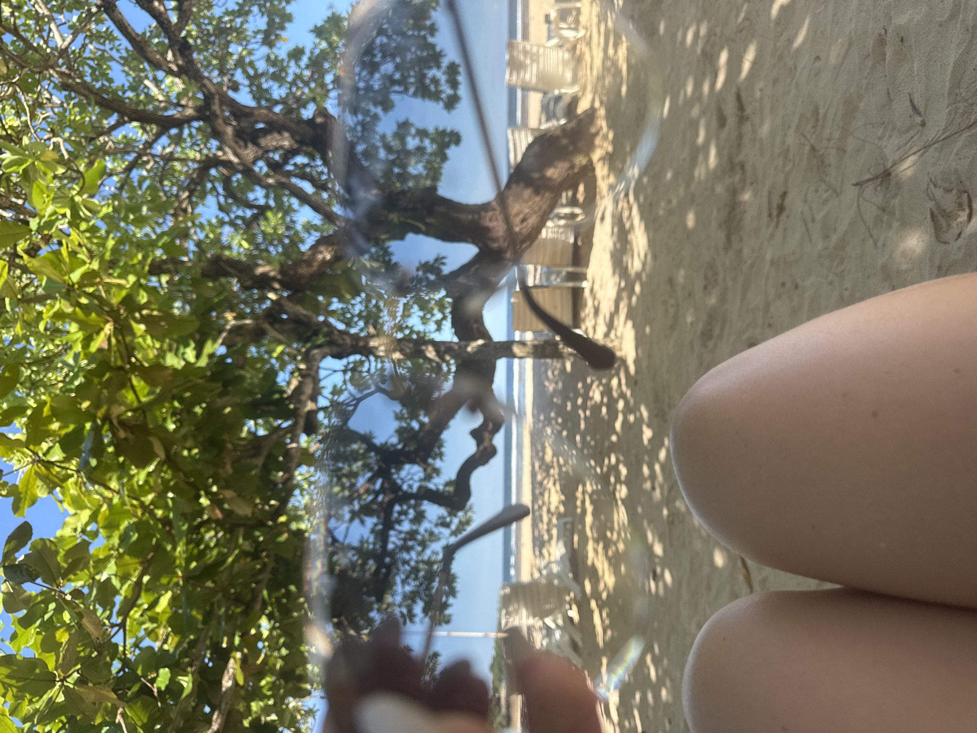 The image appears to be a photograph taken from a low angle at a beach in Kuta Selatan, Indonesia. The perspective is from someone sitting on the sand, with the person's thighs visible in the lower right corner. The focus is on a pair of sunglasses held up, reflecting the beach scene. The reflection shows a tree with green foliage to the left, and the beach itself with white chairs and the ocean in the background. The sand is light tan in color with shadows cast by trees. The sky is visible above the trees and through the sunglasses reflection, and is a clear blue. The sunglasses are partially visible, held in front of the scene. The overall impression is a casual beach setting.