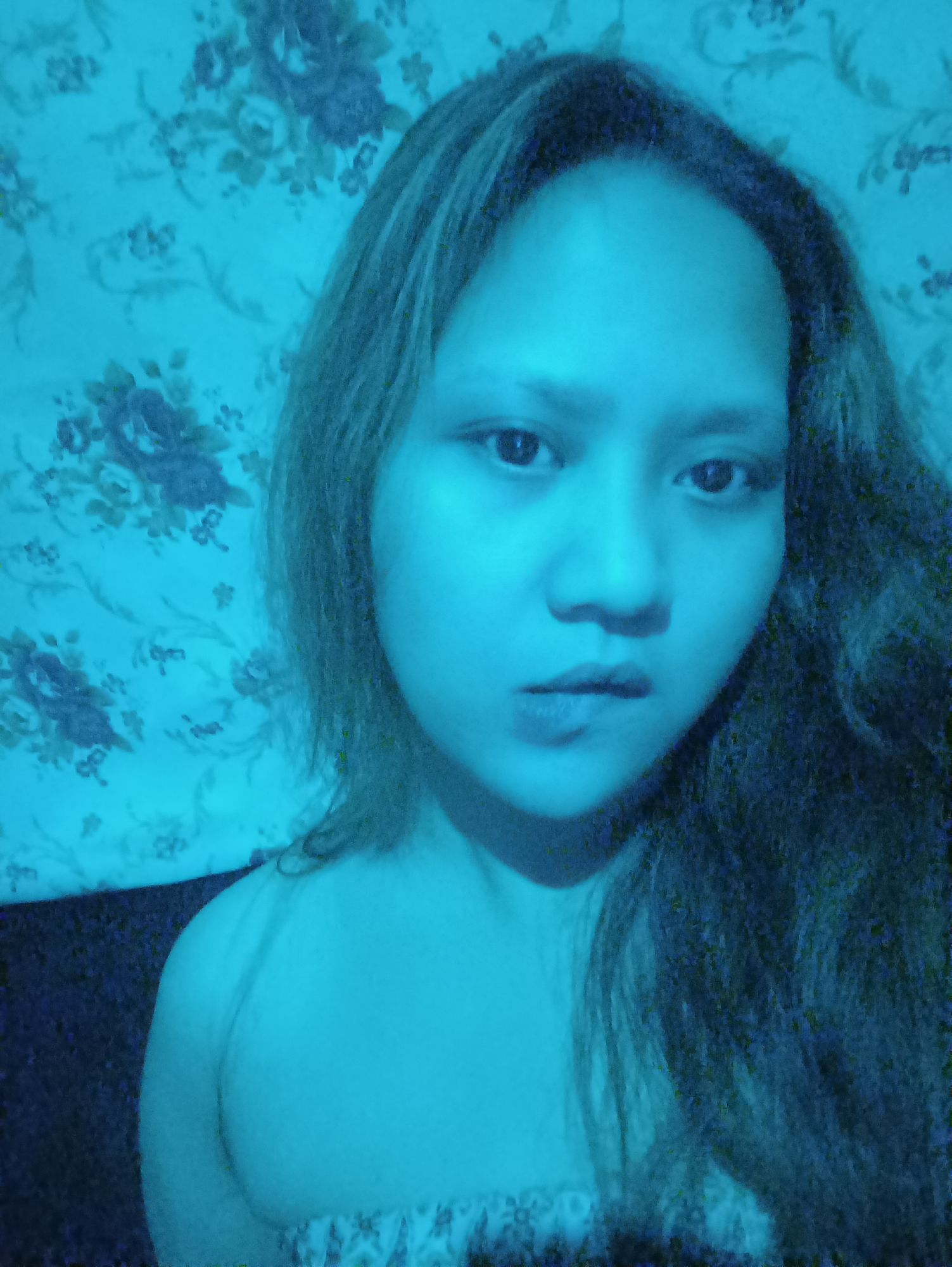 The image shows a close-up, waist-up portrait of a young woman illuminated by blue light, against a floral-patterned background that appears to be wallpaper. The woman has dark hair that is long and somewhat messy, framing her face. Her eyes are open and looking towards the camera, and her lips are slightly parted. She is wearing a strapless garment that is visible at the bottom of the frame, with a floral pattern. The overall lighting creates a cool, monochromatic tone. The scene is likely an indoor setting, possibly a bedroom or living area in a private residence in Depok, Indonesia, given the common use of floral wallpaper in homes in the region.
