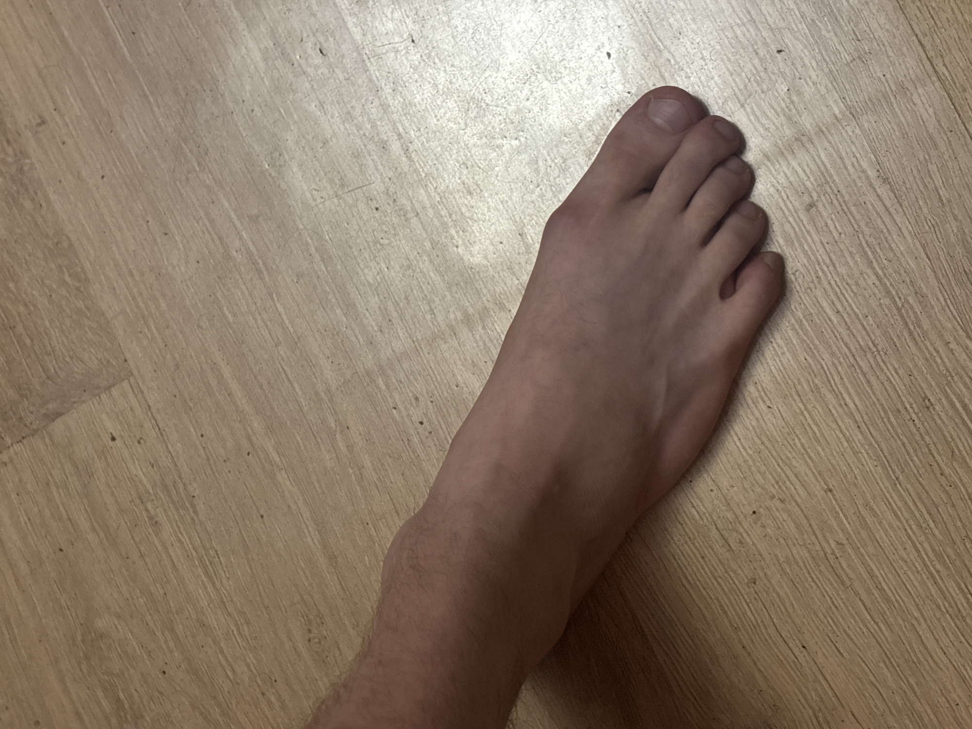 A left human foot is positioned on a light-colored wooden floor. The foot is bare and appears to be that of an adult male, with visible veins and light hair on the ankle and lower leg. The toes are spread slightly, and the toenails are visible. The wooden floor is made of planks with a visible grain pattern and appears to be slightly dusty. The lighting suggests an indoor setting. There are no other individuals or objects in the frame. The image does not provide any information to determine its location in the city of Khimki, Russia.