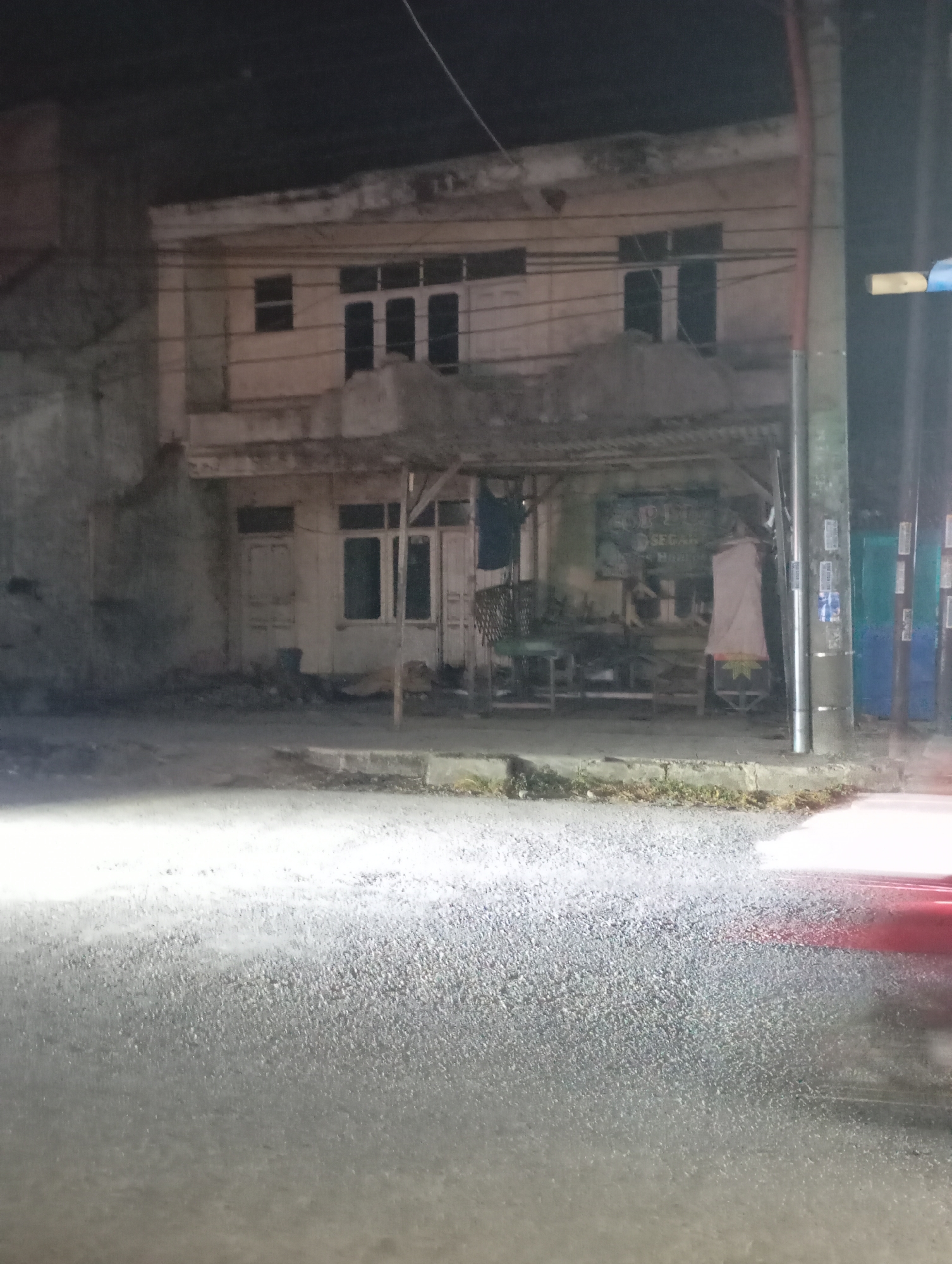 The image shows a street scene at night in front of a two-story building in Kecamatan Haurgeulis, Indonesia. The building appears to be a residence with a shop or stall at the ground level. The street is asphalt and illuminated by artificial lights, with a blurry red light streak suggesting moving traffic. The building's facade is light-colored and shows signs of wear. The ground floor has several windows and doors, and a covered area in front of it serves as a stall, displaying some items and what appears to be a chair. Electrical wires are visible above the building. The overall environment is dimly lit and somewhat cluttered.