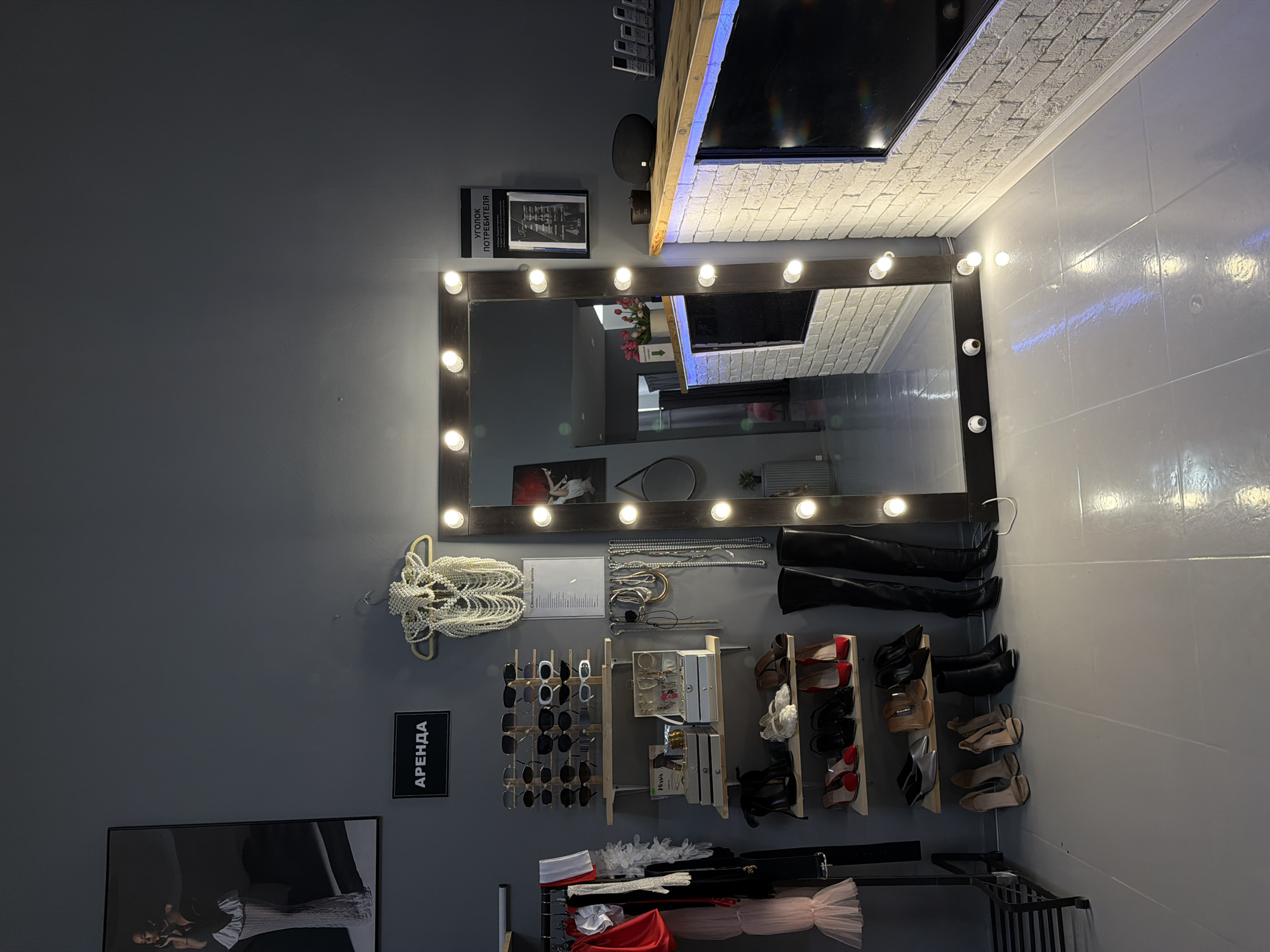 This appears to be a dressing or styling room, possibly in Kazan, Russia. The room has a large, illuminated mirror surrounded by light bulbs. Beneath the mirror, there are shelves and racks displaying various items including: accessories such as pearl necklaces, jewelry, sunglasses, a pair of over-the-knee boots, and shoes. Clothing, including tutus, is visible on racks. A sign that reads "APEHДA" (which translates to "RENT") is located near a black and white poster with a picture of a person. Additionally, a sign above the mirror which translates to "customer's corner" in English. The walls are painted in shades of grey.