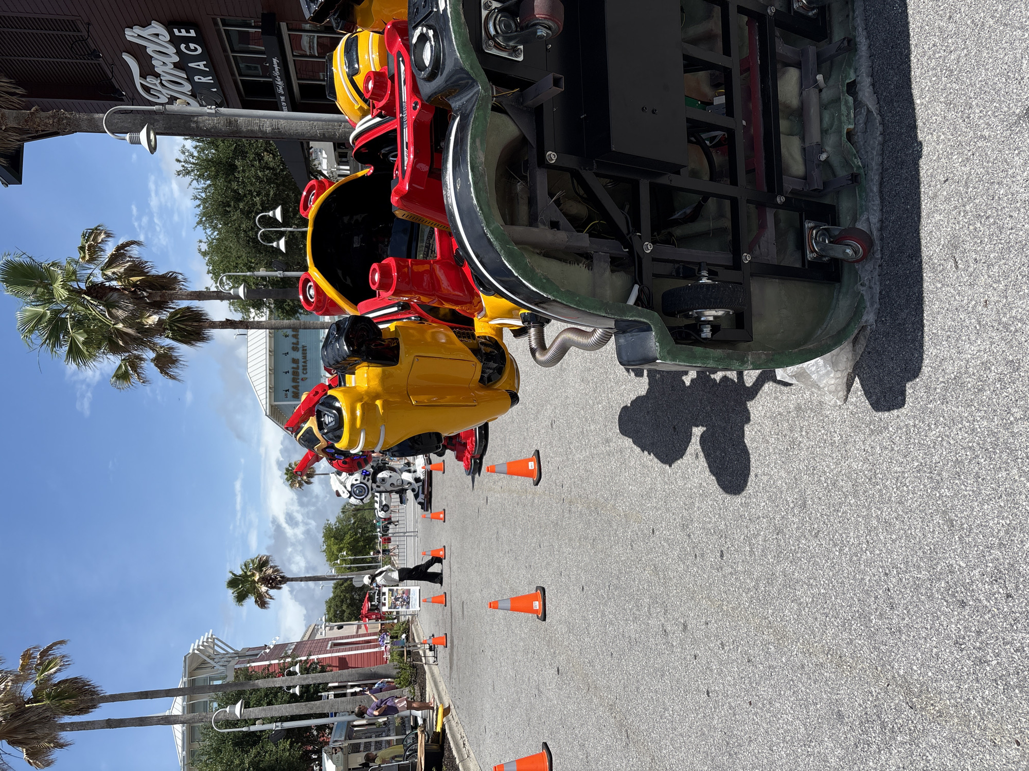The image shows a street in Panama City Beach, Florida, United States, lined with novelty vehicles resembling robots or machines. The vehicles are painted in bright colors like yellow, red, and black. The street is partially blocked off by orange traffic cones. A man in dark clothing is visible between the vehicles. Palm trees and building facades are visible in the background, one with the sign "Ford's Garage" on it. The sky is blue with scattered clouds, indicating a sunny day. People are visible near the sidewalk.