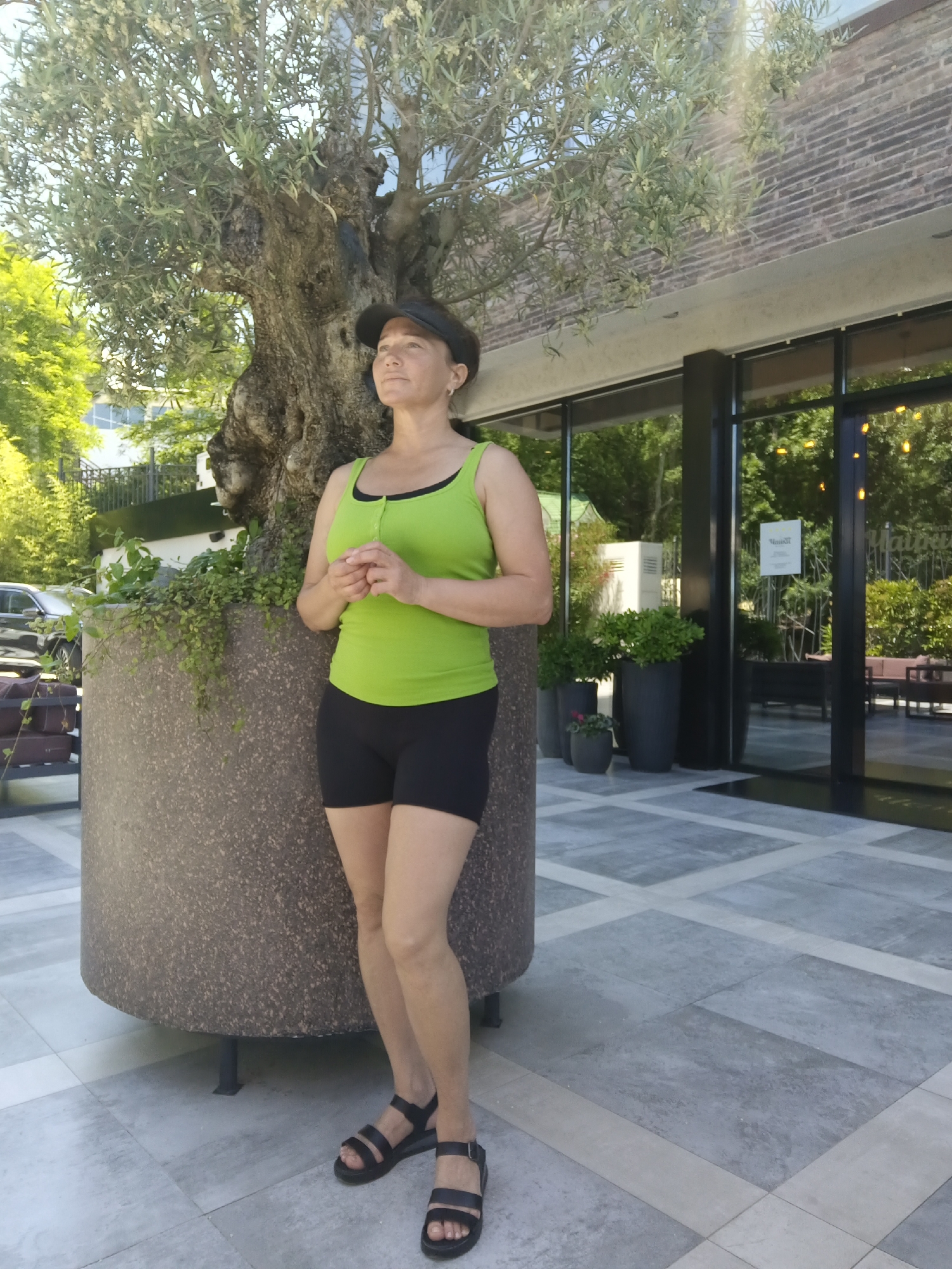 A medium shot captures a woman standing near a large, textured planter and an olive tree. She is wearing a lime green tank top and black shorts, with black sandals on her feet and a dark visor on her head. Her hands are clasped in front of her. She is looking upwards and to her right, with her head tilted slightly. In the background, a building with large windows and a brick facade is visible, along with greenery and outdoor seating areas. The ground is paved with large, grey tiles. The scene appears to be outdoors in an urban setting, possibly a restaurant or hotel patio.