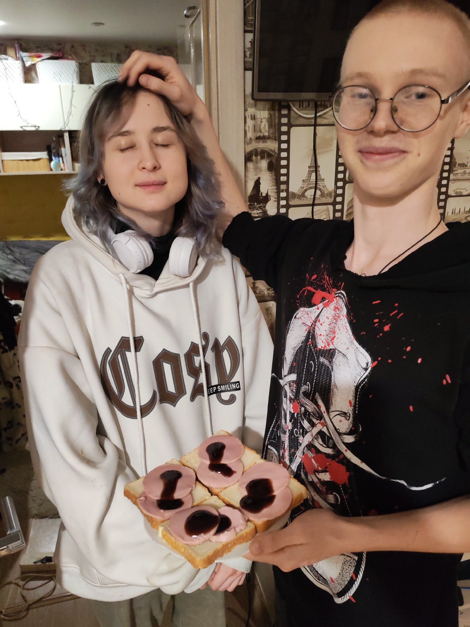 Two individuals are indoors, positioned near a door and a wall adorned with a film-themed wallpaper. The person on the left is wearing a white hoodie with the word "Cozy" printed on it, and headphones are draped around their neck. Their eyes are closed, and a hand is touching their head. The person on the right is wearing a black hoodie with a graphic design and eyeglasses. They are holding a plate with four open-faced sandwiches, each topped with slices of meat and a dark sauce. The location is Chaikovsky, Russia.